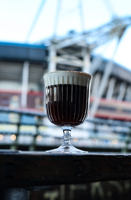 A glass of dark coffee with a layer of foam, set on a wooden surface with a modern building and blue sky in the background.