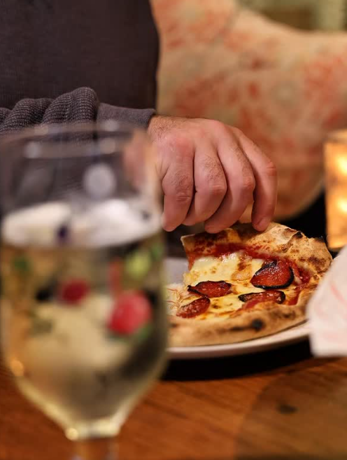 Close-up of a hand reaching for a slice of pepperoni pizza on a plate, with a glass of water and a blurred background including a patterned pillow and warm lighting.