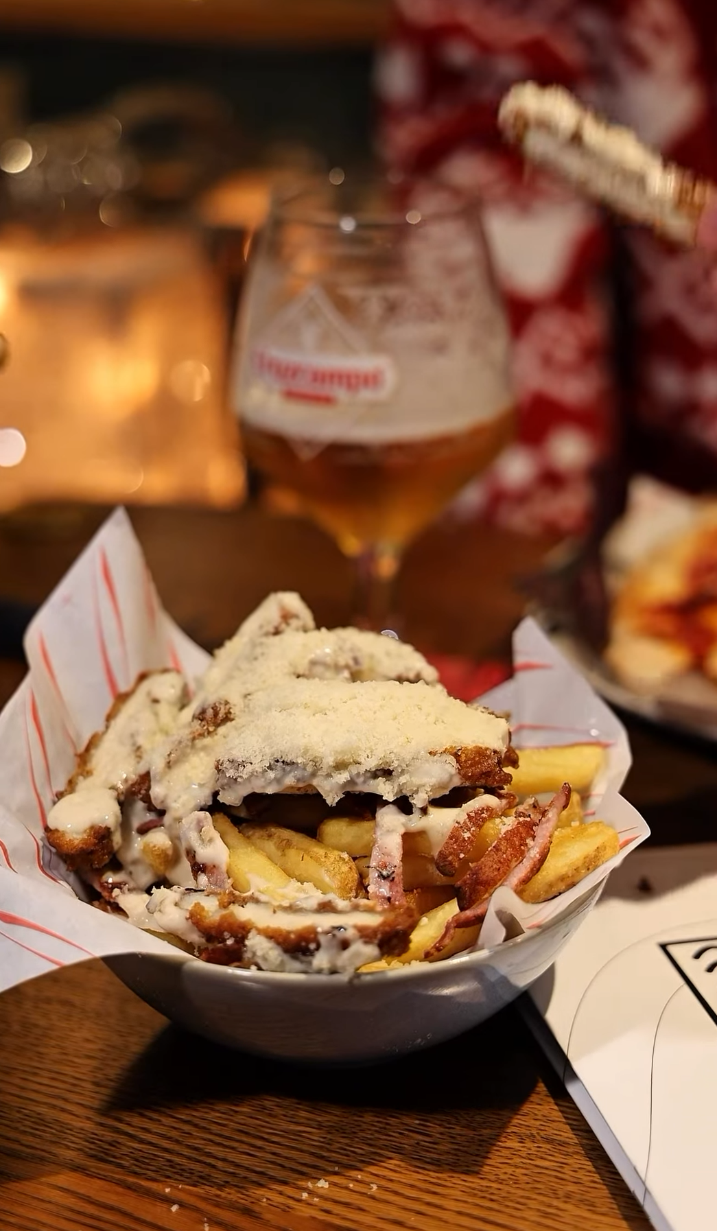 A bowl of poutine with French fries, cheese curds, gravy, and bacon bits, topped with a fried chicken fillet and a sprinkle of cheese powder. In the background, a glass of beer and a person wearing a red and white patterned shirt are visible.