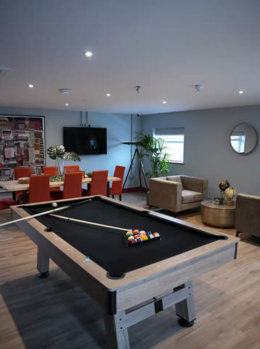 A modern lounge area with a pool table, a dining table with orange chairs, a wall-mounted TV, and seating with decorative pillows, in a well-lit room with hardwood floors and a large window.