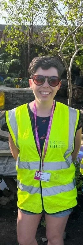 A woman with short dark hair wearing sunglasses, a bright yellow safety vest over a dark shirt, and a pink lanyard, standing outdoors with green trees and a stone wall in the background on a sunny day.