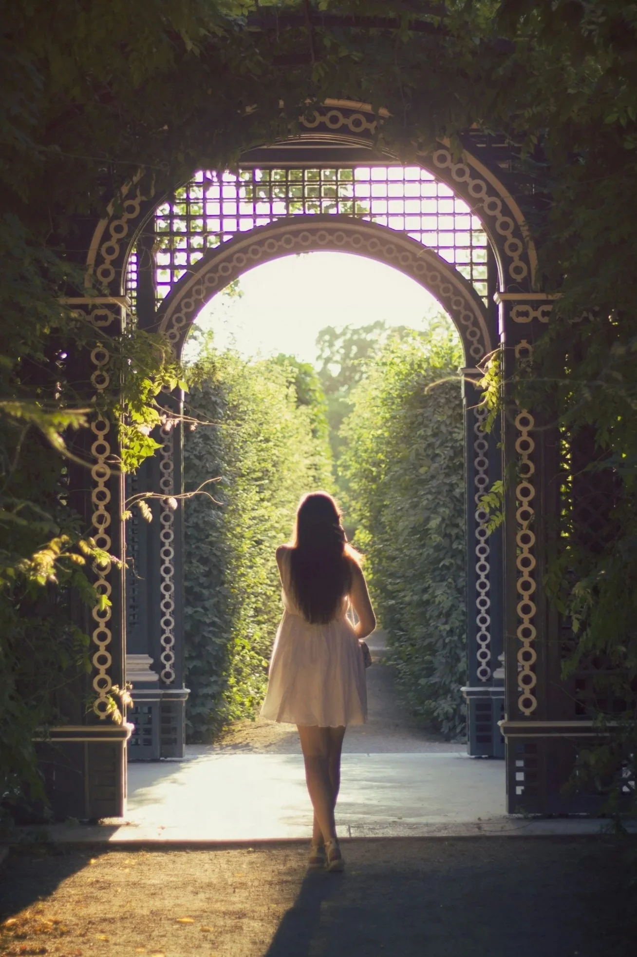 A woman in a white dress walking through a garden archway into a green, leafy path with sunlight filtering through.