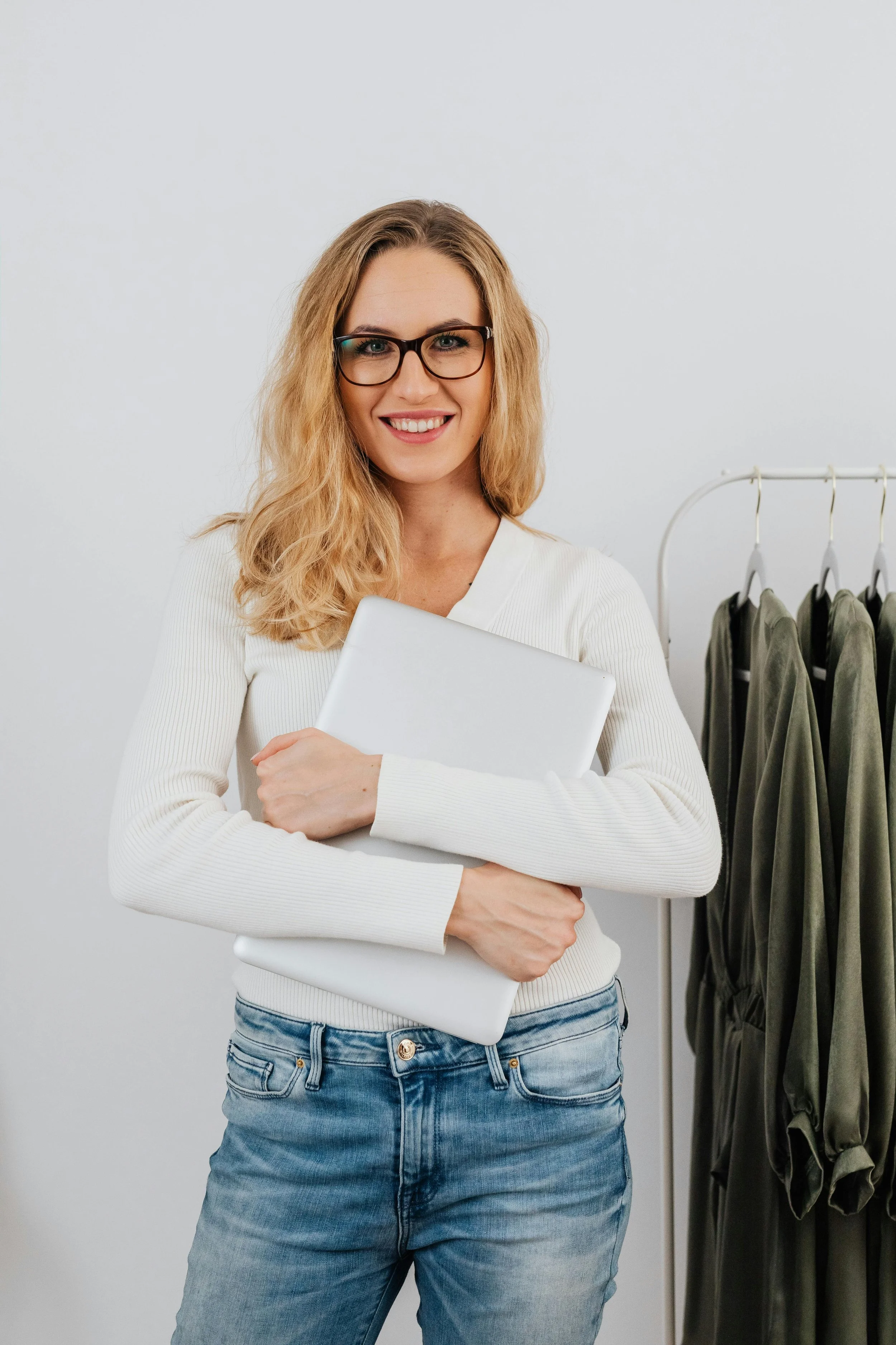 A woman with blonde hair, glasses, and a white sweater holding a closed laptop, standing in front of a clothing rack with green garments.