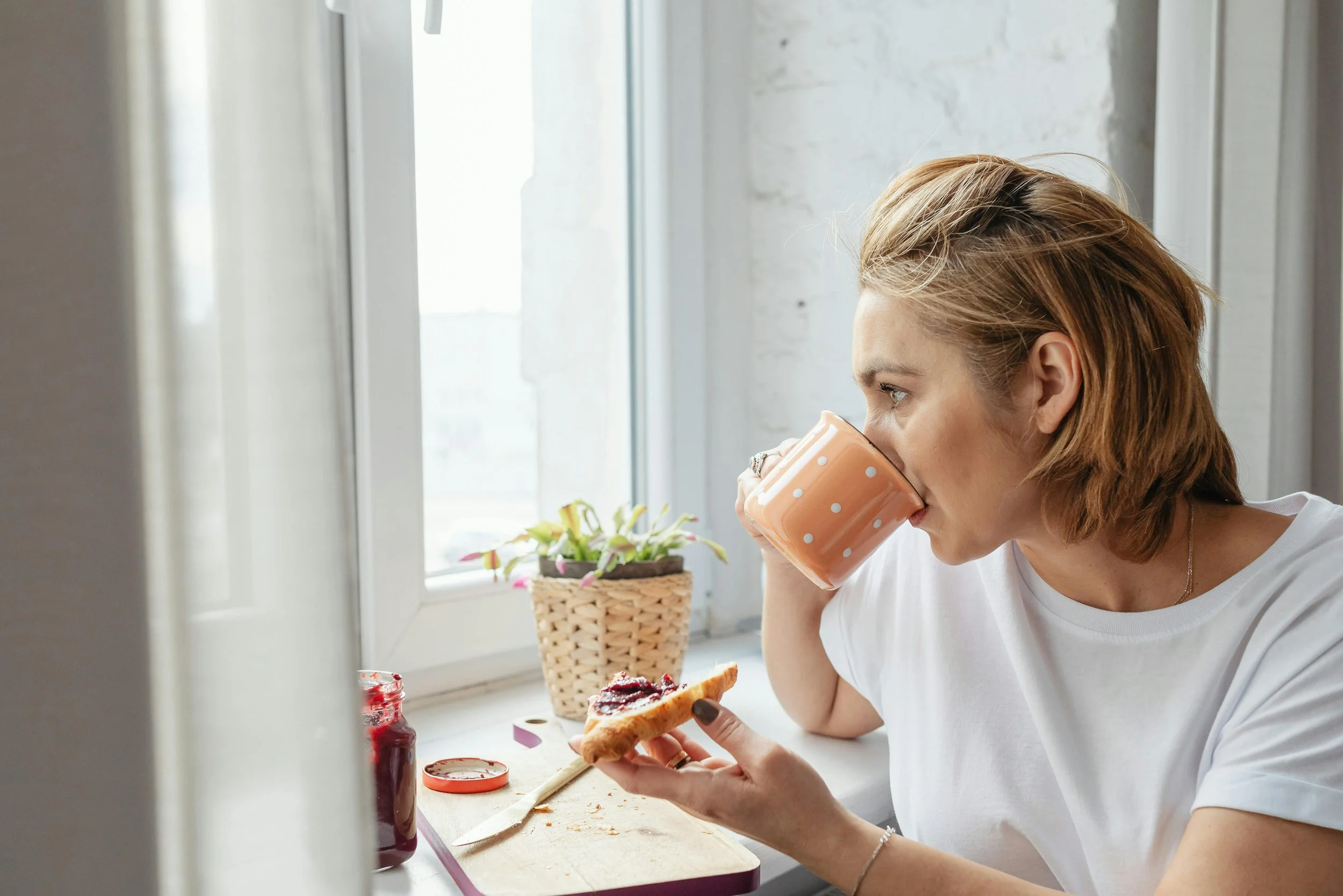 A woman with light brown hair drinking coffee while holding a slice of toast with jam near a window.