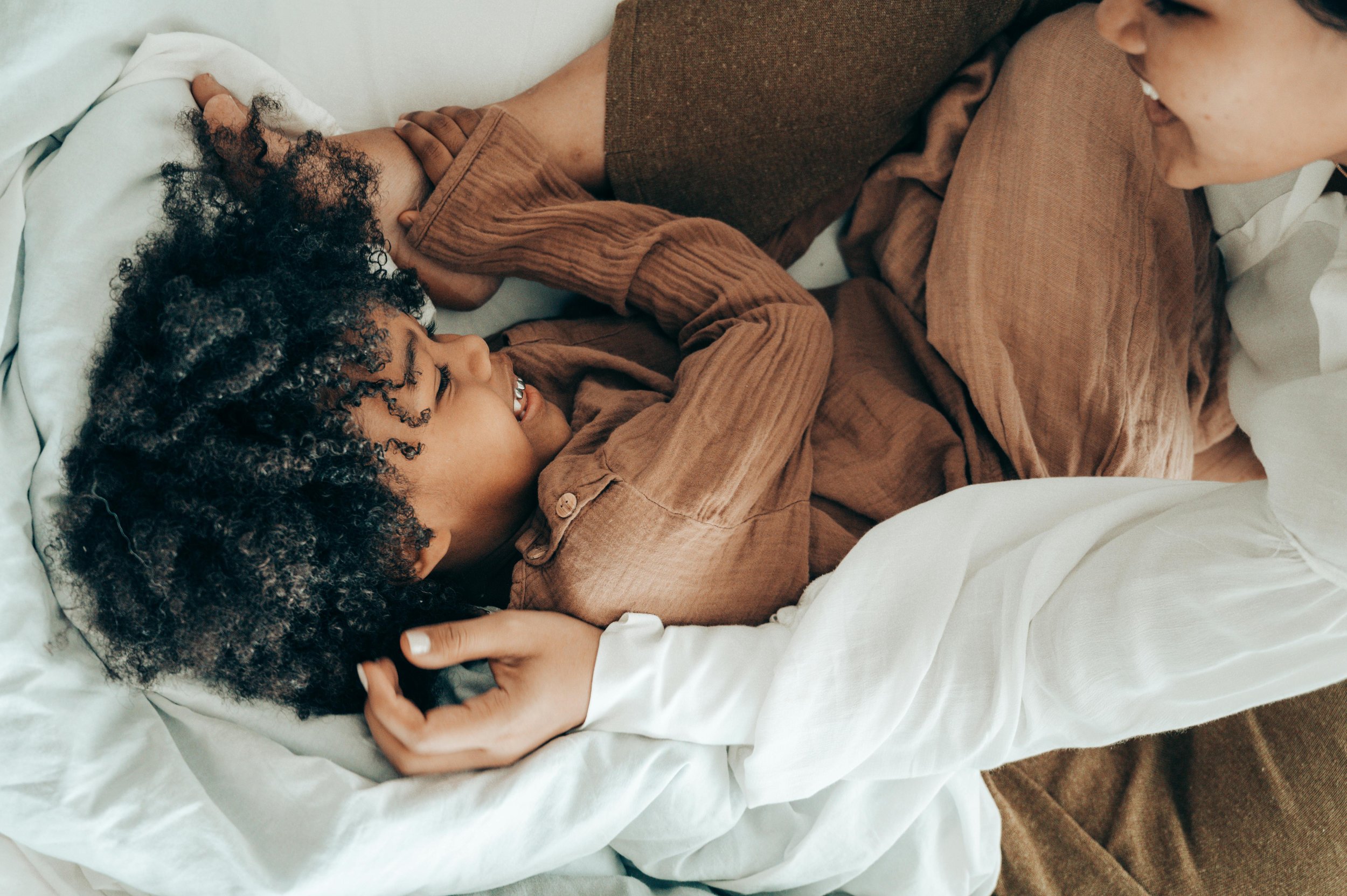 A woman and a young girl are lying on a bed, smiling and playing together. The girl has curly black hair and is wearing a brown shirt, while the woman has short hair, a light smile, and is dressed in a light-colored top. They are holding hands and sharing a joyful moment.