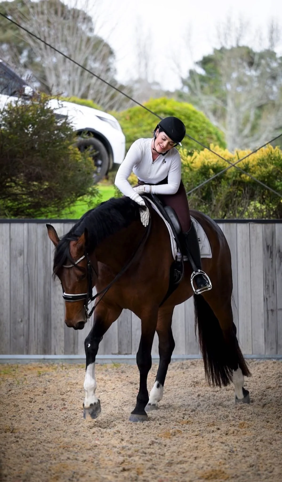 A woman wearing a black riding helmet and white riding jacket is smiling while riding a brown and white horse in an outdoor riding arena.