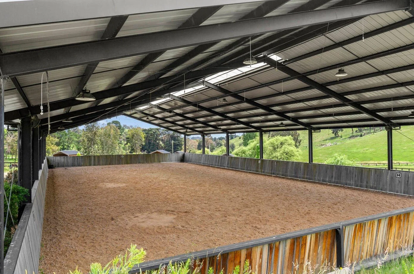 Indoor horse riding arena with sandy floor, metal roof, and open sides revealing green fields and trees outside.