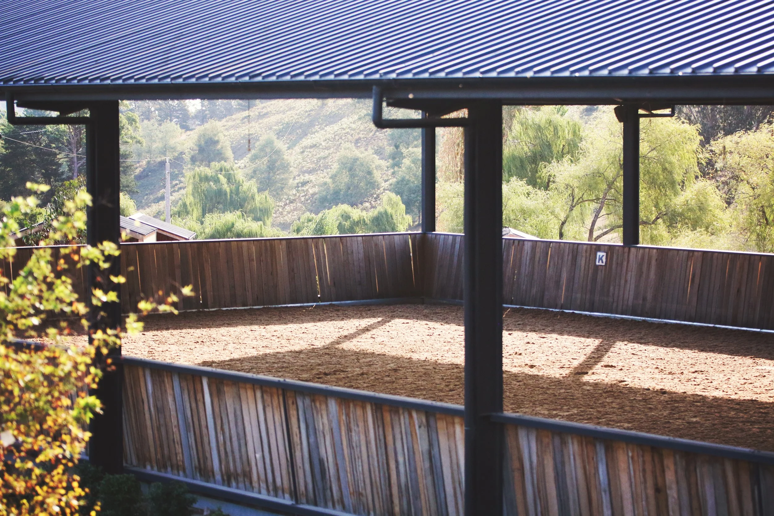 Empty outdoor riding arena with a wooden fence, surrounded by trees and hills in the background.