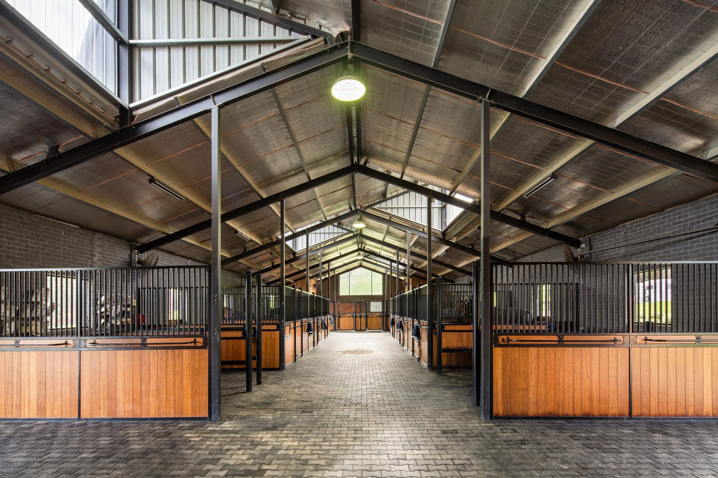 Interior of a horse stable with wooden stalls on either side and a paved walkway in the center.