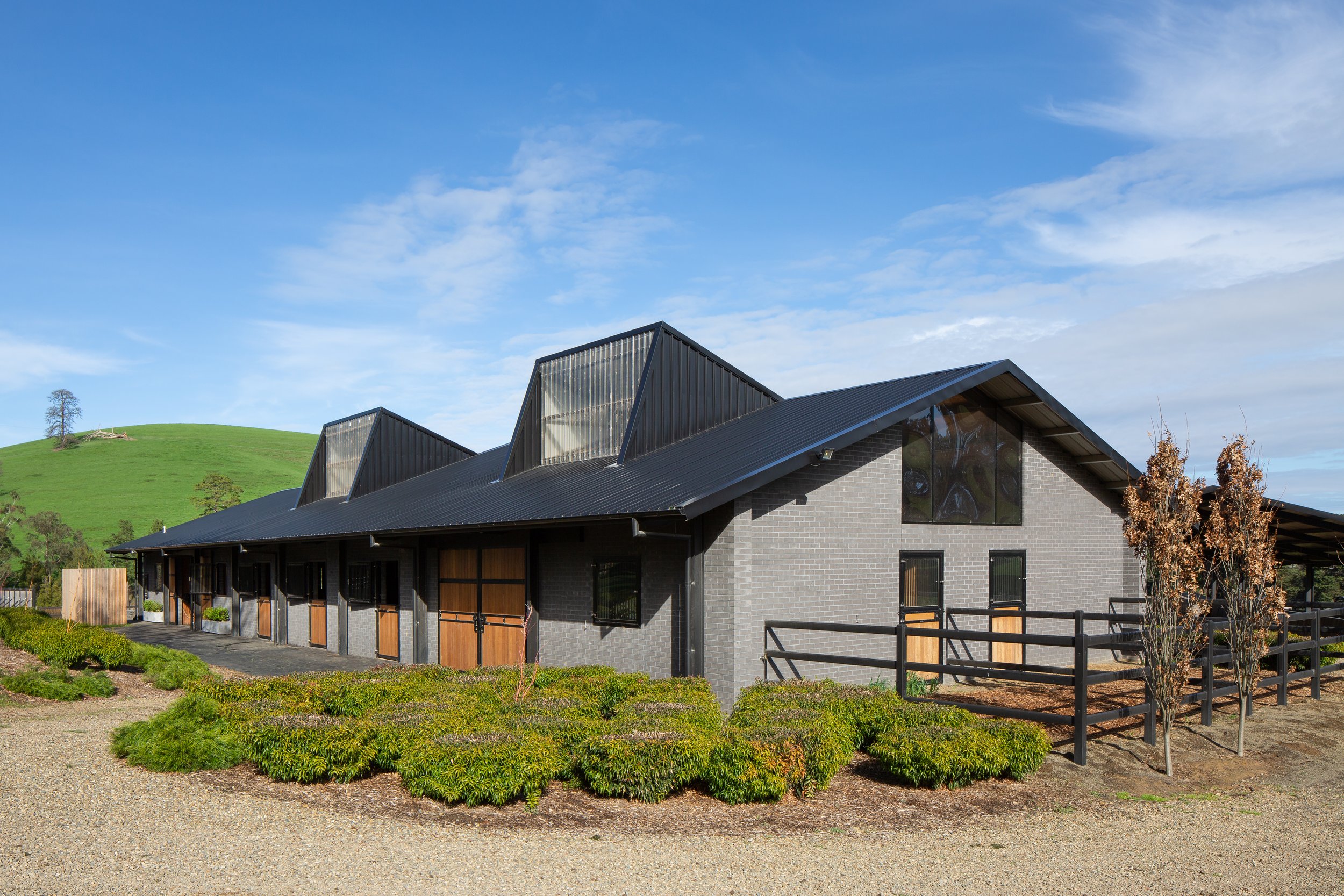 A modern building with a black metal roof and gray brick walls, featuring large triangular roof windows. Surrounding greenery and a gravel pathway are visible, with a grassy hill and trees in the background under a partly cloudy sky.