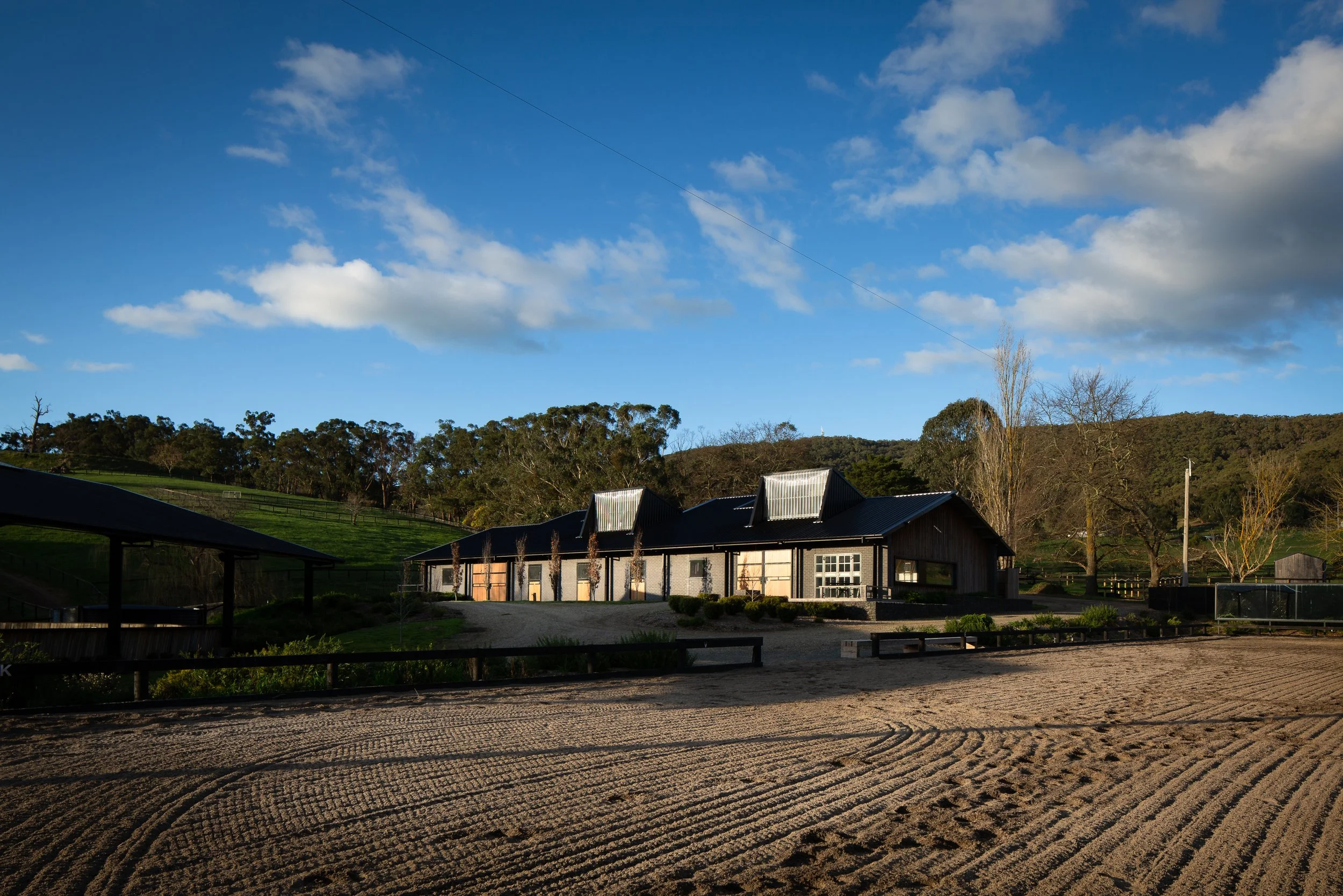 A rural landscape with a modern house, dirt foreground, green hills, trees, and a partly cloudy blue sky.