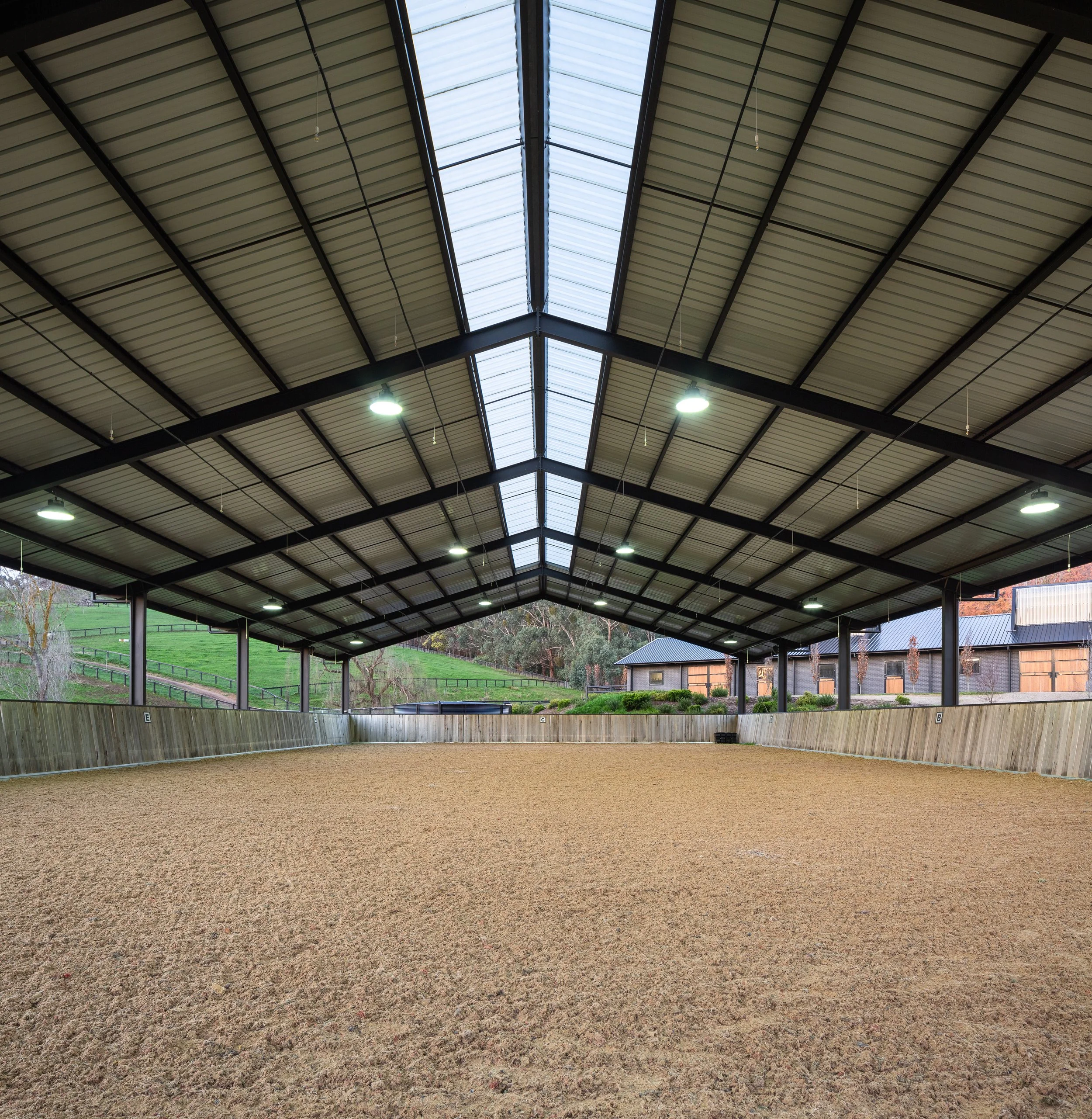 Indoor equestrian riding arena with a sandy floor, wooden sidewalls, and a high metal roof with skylights and hanging lights.