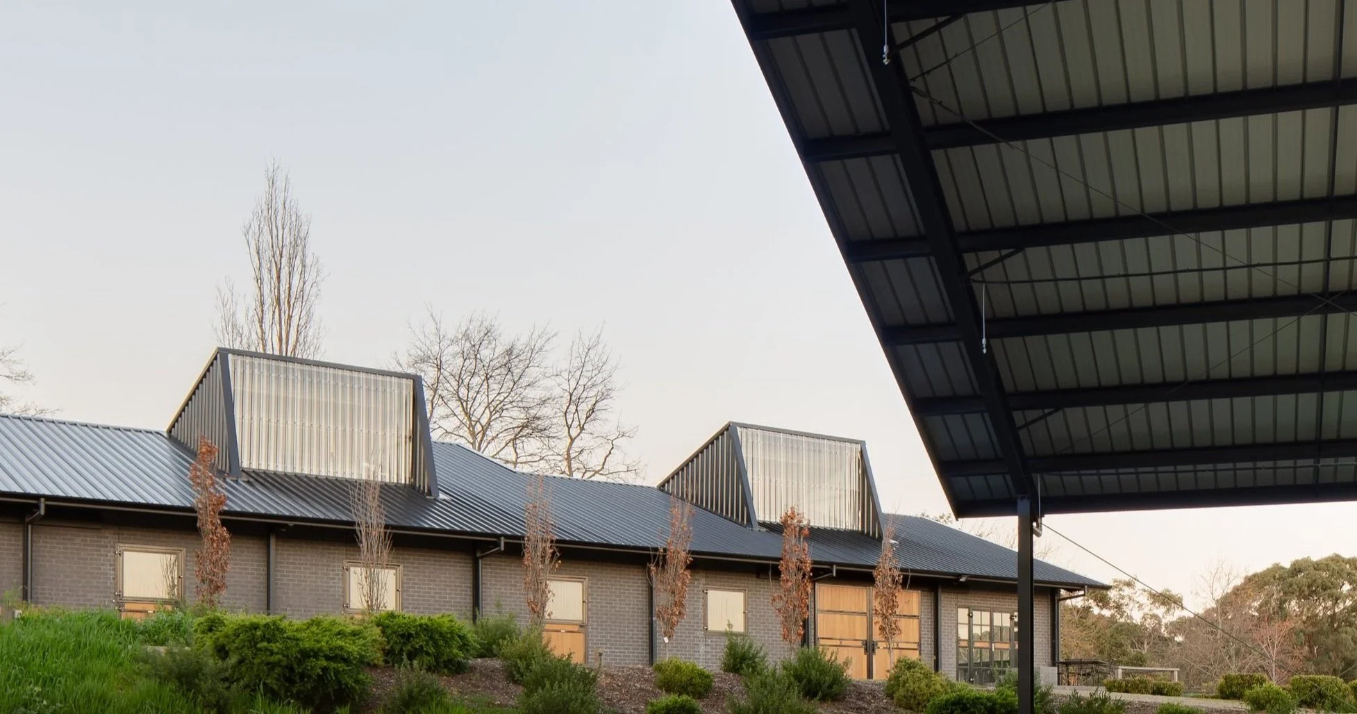 A modern building with metal roofing and large windows, surrounded by landscaped greenery, trees, and an outdoor covered area with a metal roof.