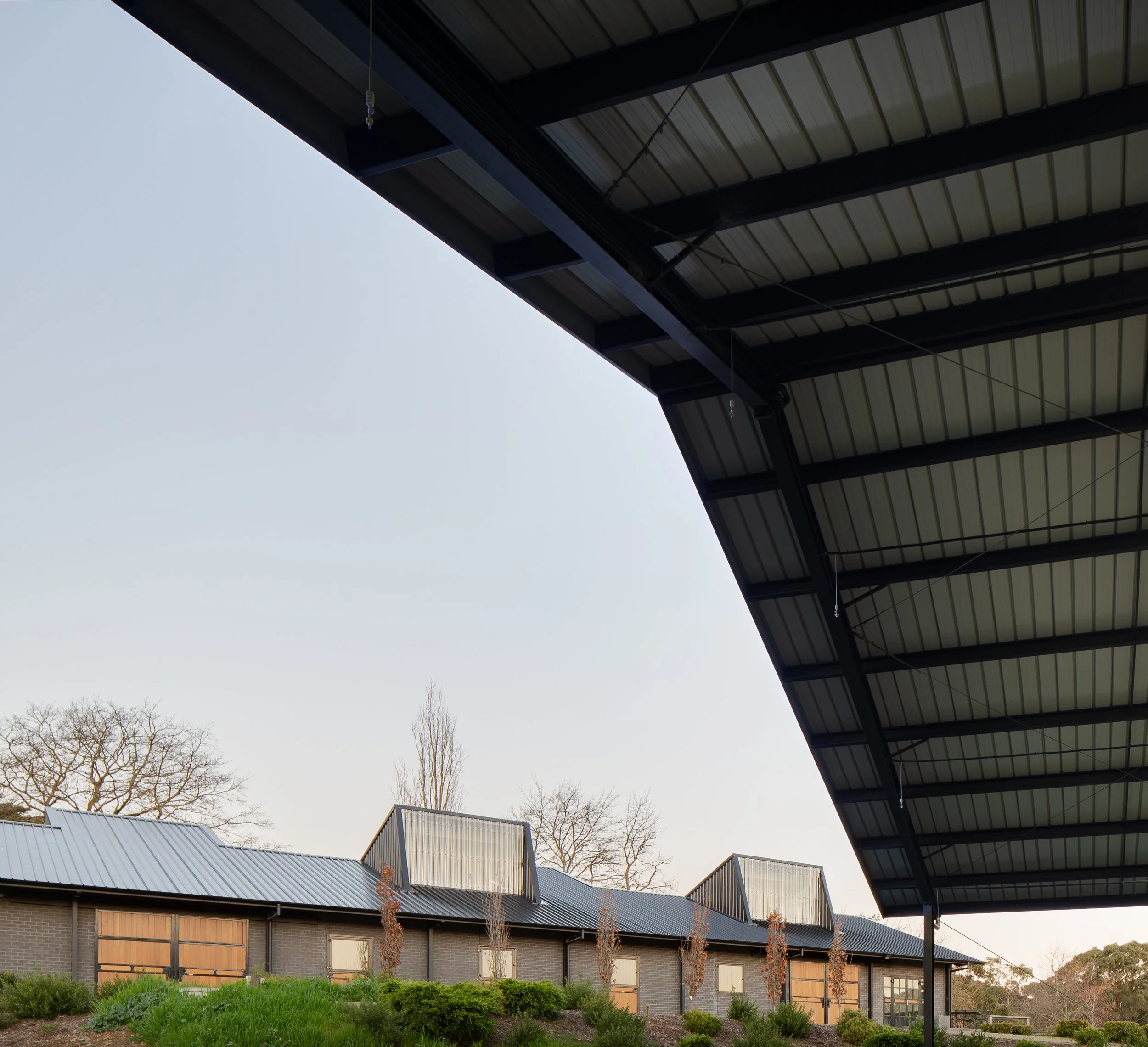 View of a row of modern townhouses with solar panels on the roofs, seen from under a large metal roof structure, with trees in the background and shrubbery in the foreground.