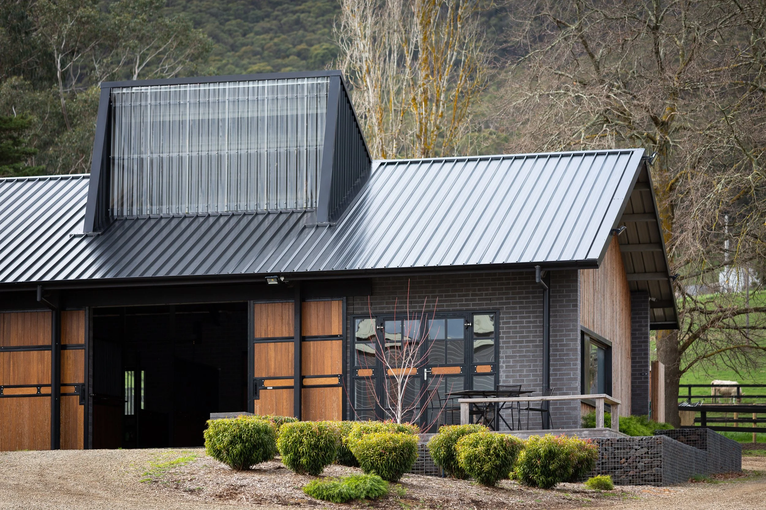 Modern house with dark brick and wooden exterior, metal roof, large window, outdoor seating, surrounded by bushes and trees