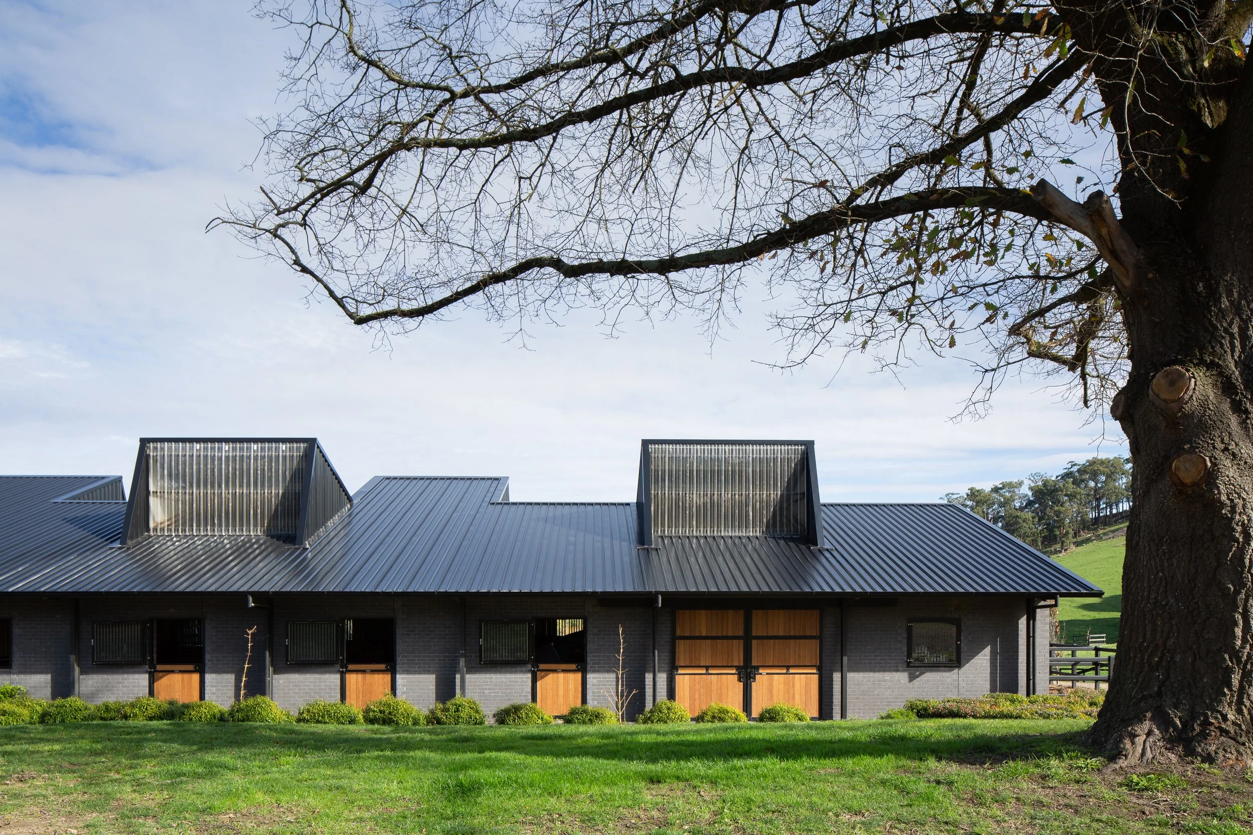 A modern gray brick barn with a black metal roof and wooden doors, situated on a grassy field with a large leafless tree in the foreground and green hills in the background.