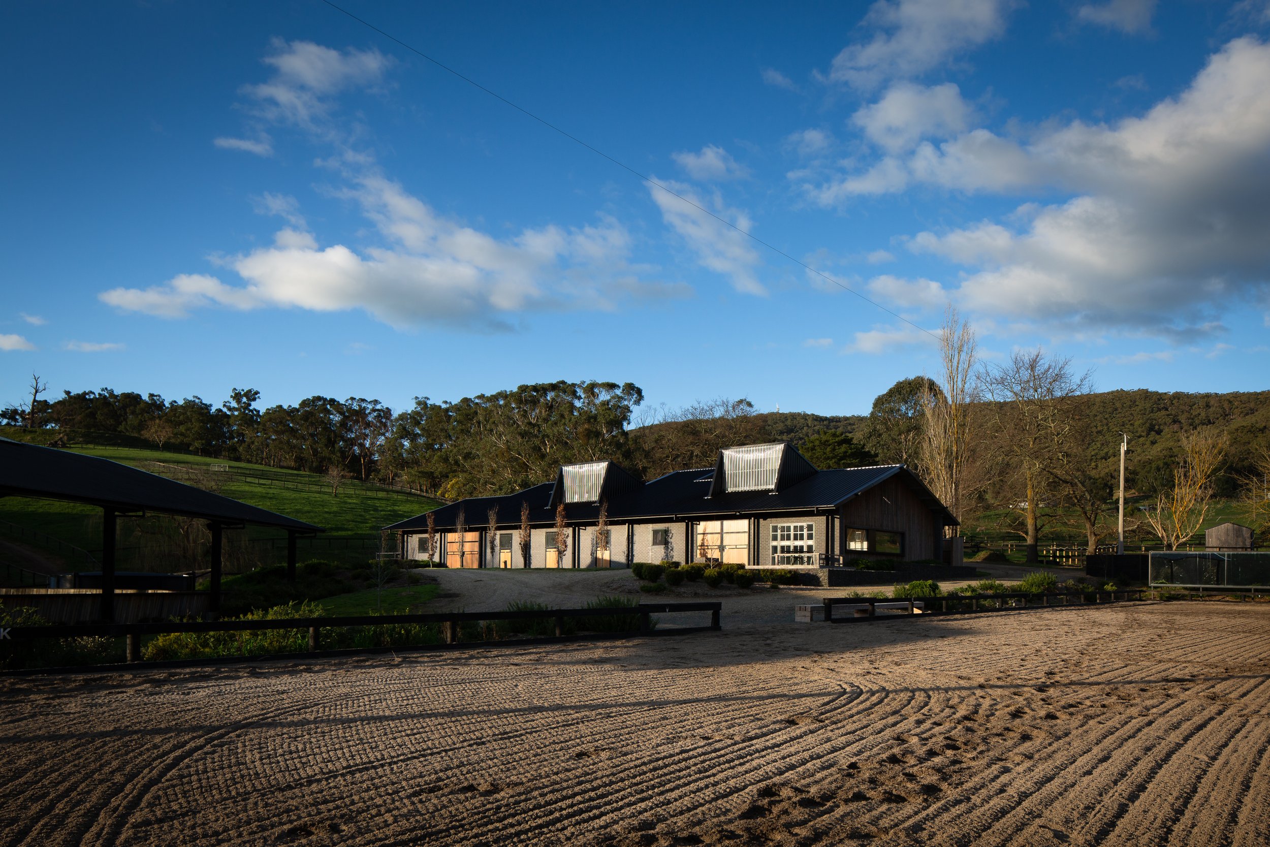 A rural farm scene with a large barn-like building, gravel driveway, and surrounding trees under a partly cloudy blue sky.