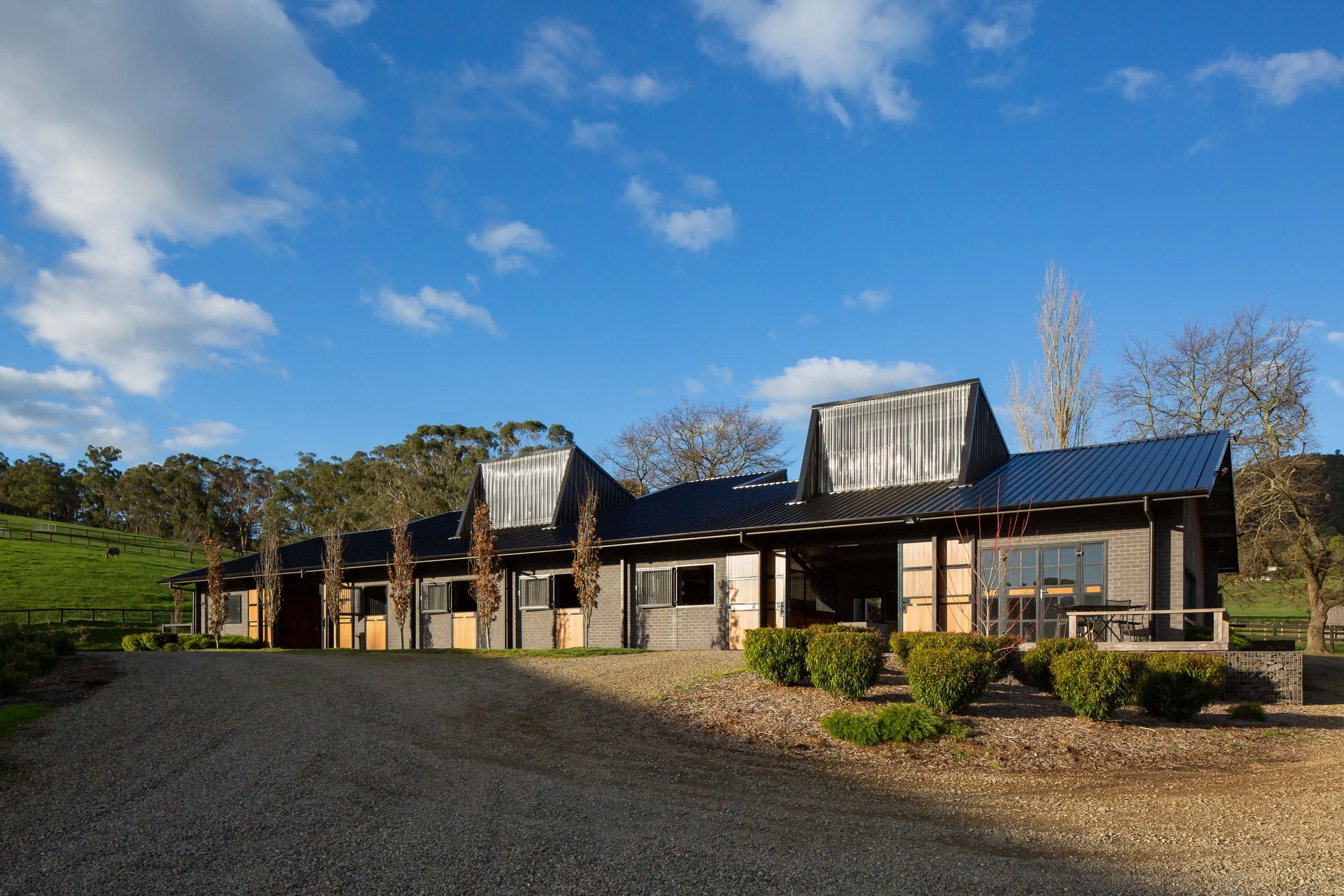 A modern farmhouse with a black metal roof, dark brick walls, and wooden accents, situated on a rural property with a gravel driveway, manicured bushes, leafless trees, and green pastures in the background under a blue sky with scattered clouds.
