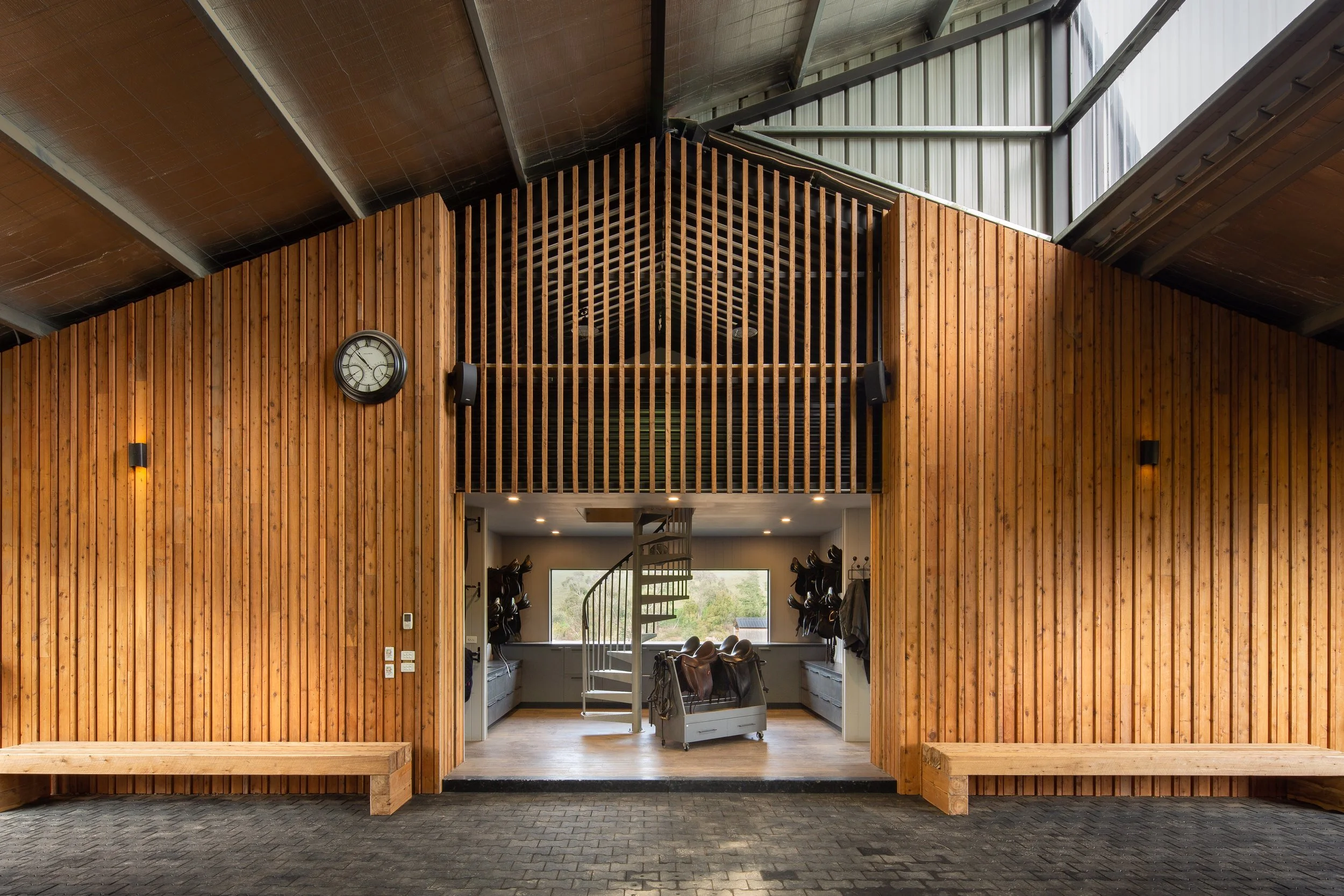 Interior of a modern riding stable or equestrian center with wood-paneled walls, a spiral staircase, benches, clocks, and riding gear hanging on walls.
