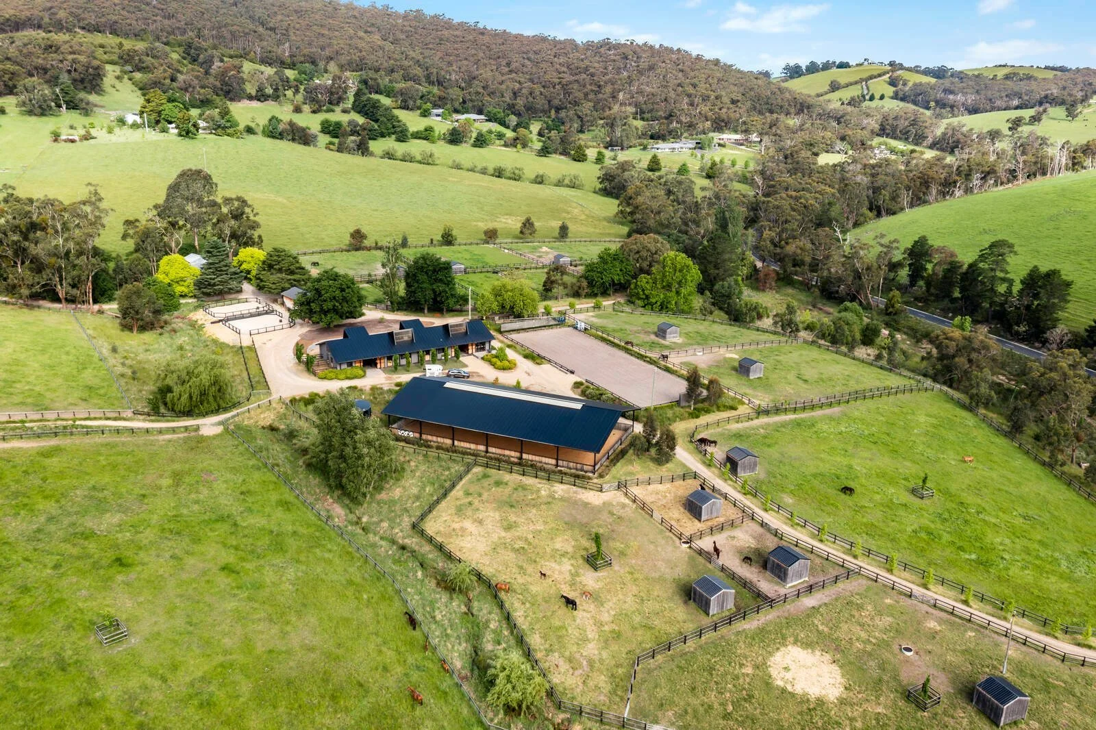 Aerial view of a horse farm with several fenced paddocks, a large barn with a dark roof, and multiple smaller structures, surrounded by rolling green hills and trees.