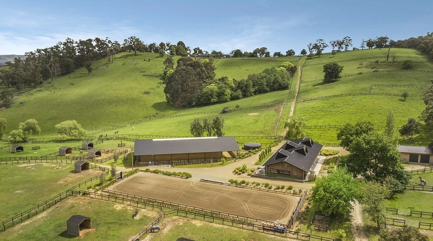 A sprawling farm with green rolling hills, several trees, a large stable with a dark roof, smaller barn structures, a dirt riding arena, and a few horses grazing on the pasture under a clear blue sky.