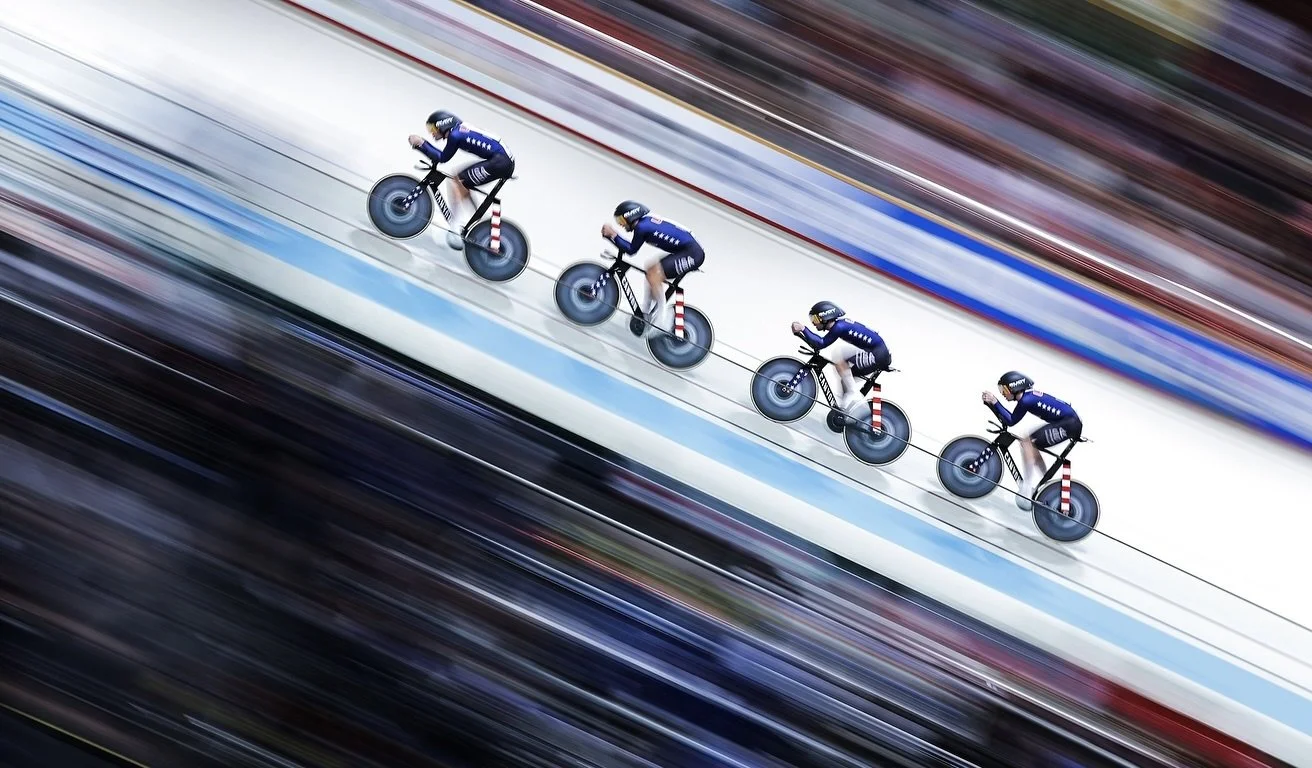 Four cyclists in a line riding on a velodrome track with blurred spectators in the background.