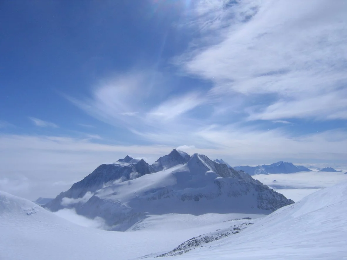 Snow-covered mountain peaks beneath a partly cloudy sky, with clouds settling in valleys.