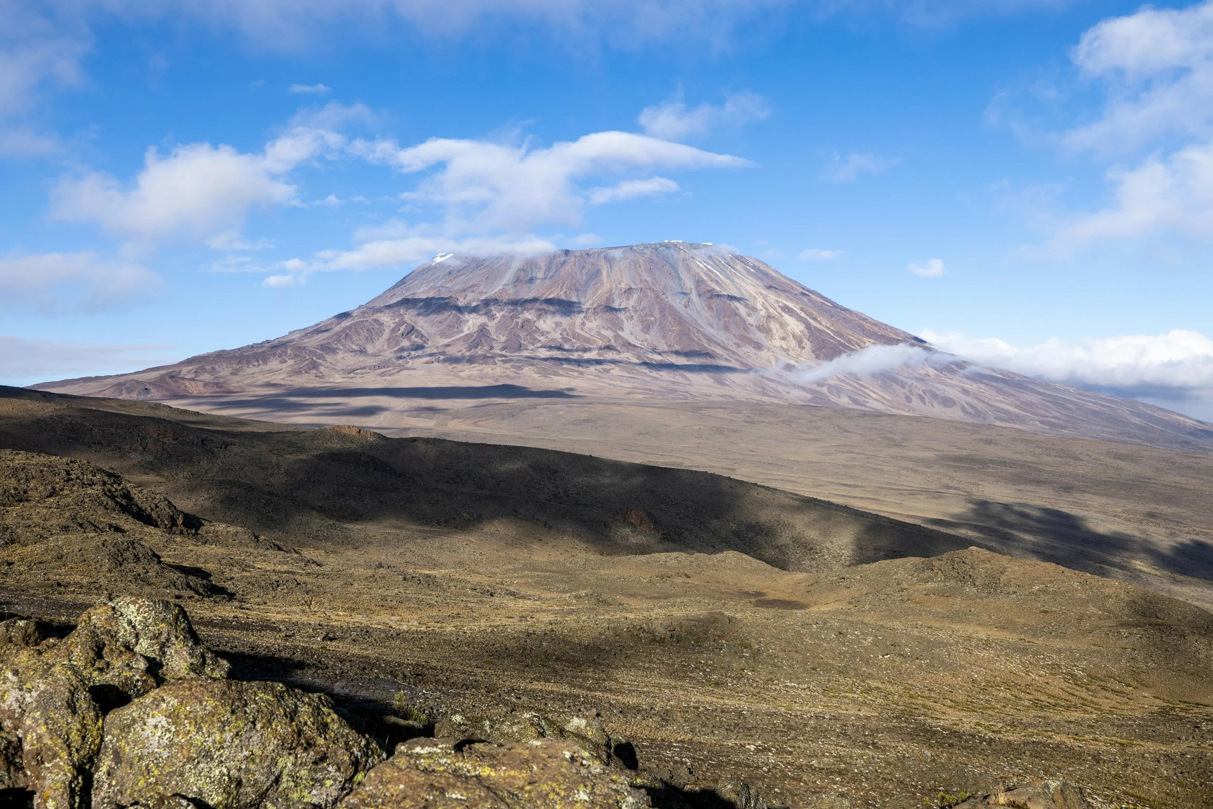 A large volcano with a snow-capped peak in the distance and a barren, rocky landscape in the foreground under a partly cloudy sky.