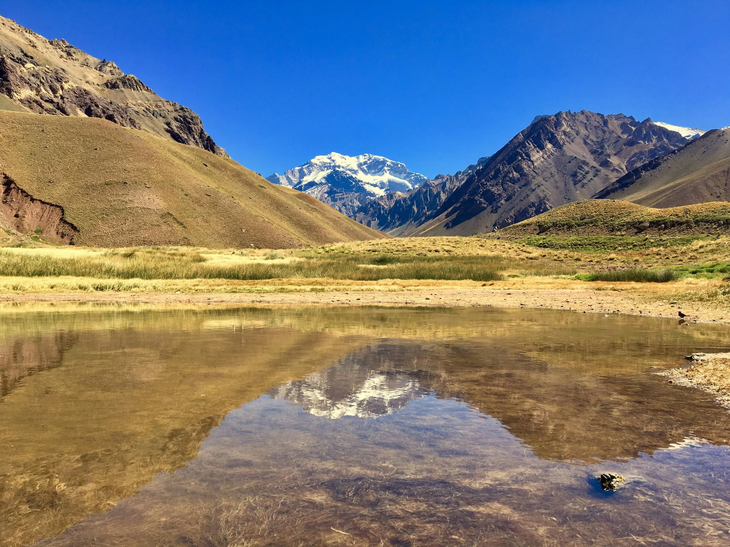 A mountain landscape with a snow-capped peak in the background, brown and green hills, and a reflective body of water in the foreground.