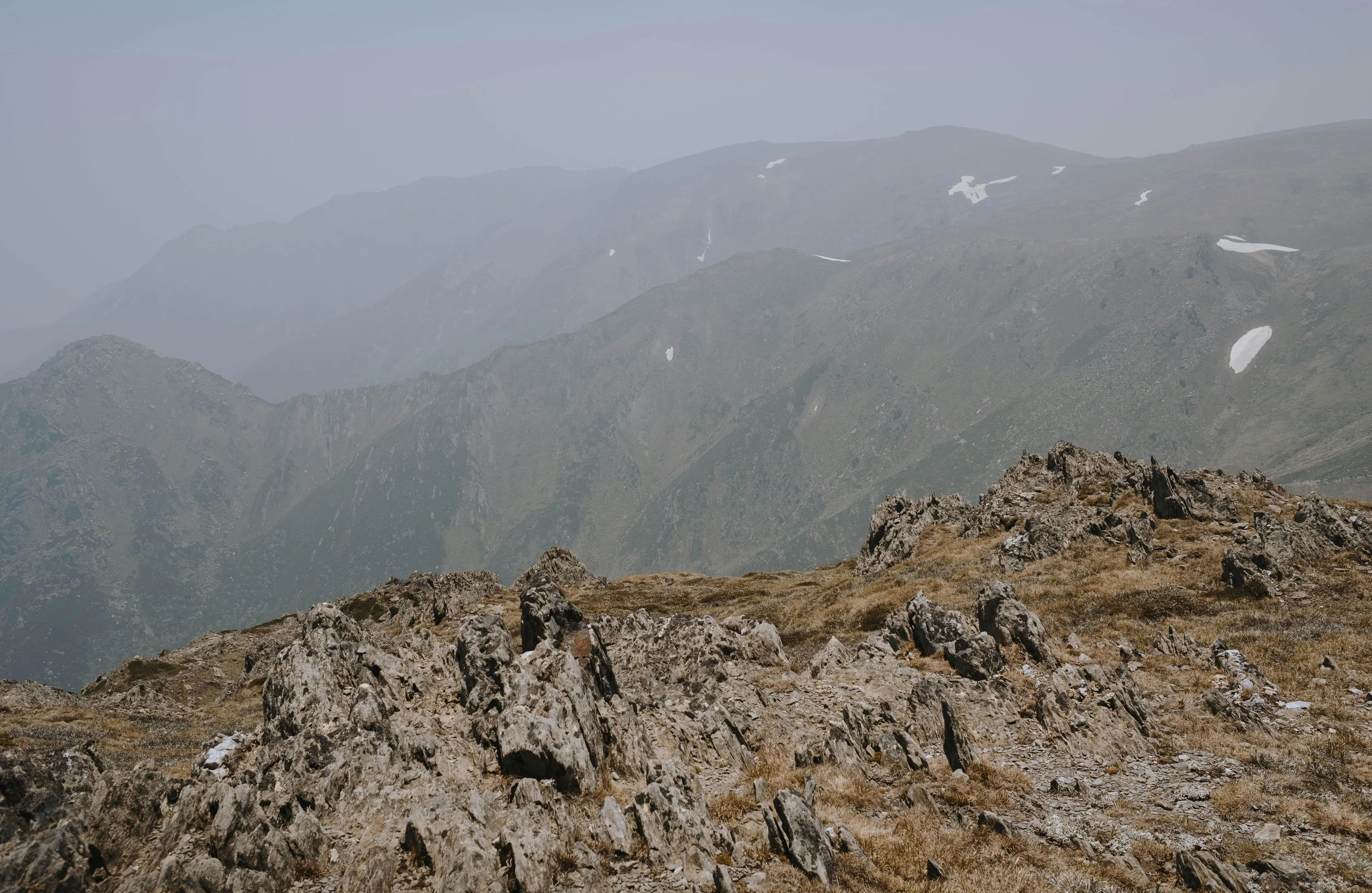Mountain landscape with rocky terrain in the foreground, green slopes and patches of snow on distant peaks under a hazy sky.
