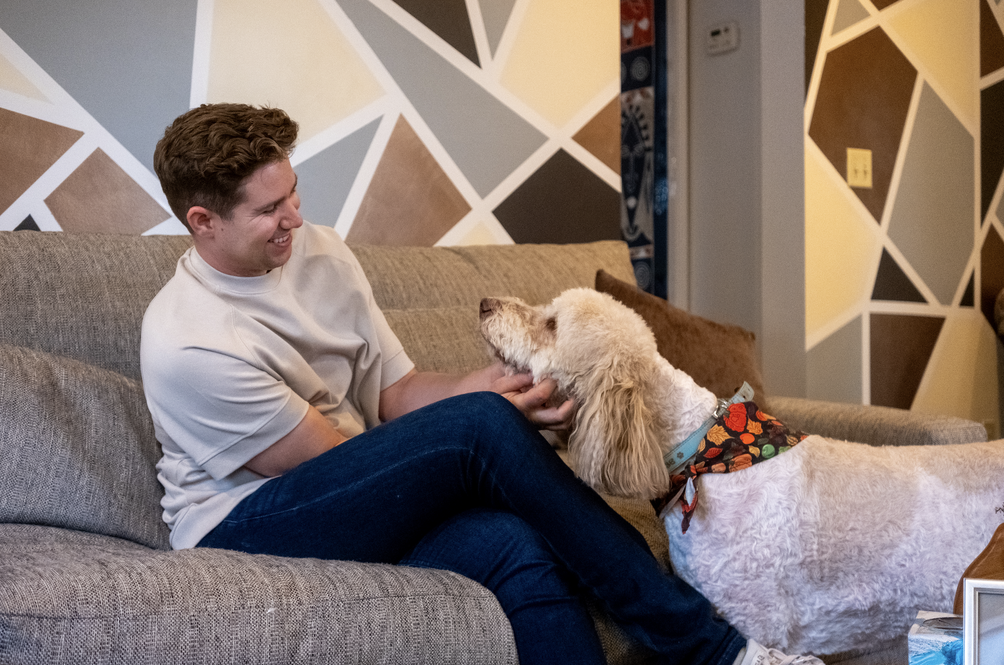 A young man sitting on a gray couch smiling as he interacts with a large, light-colored dog with curly fur. The dog is wearing a colorful bandana and looking up at the man.