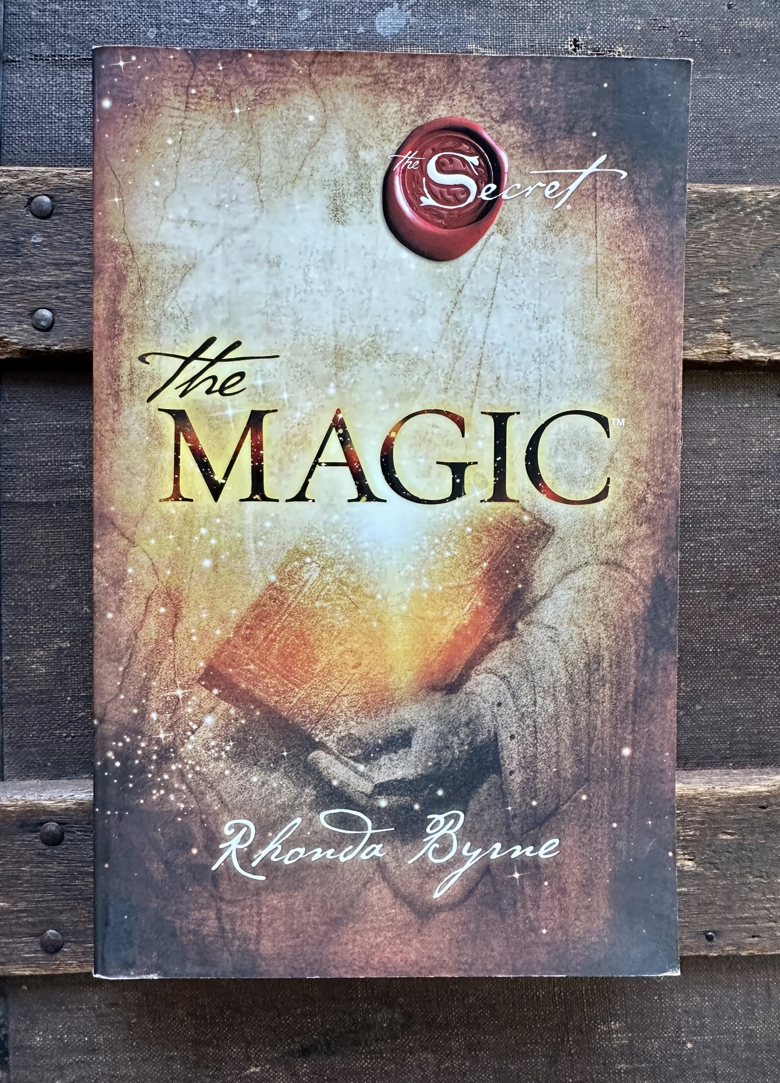 Book cover titled "The Magic" by Rhonda Byrne featuring a mystical glowing open book emitting sparkles, with a background resembling a textured, cosmic surface.