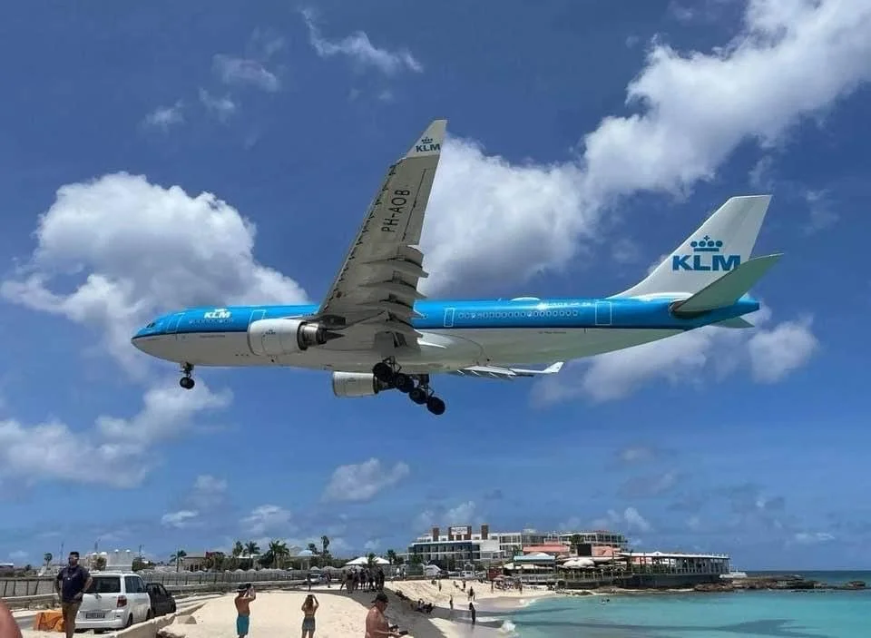 A KLM airplane flying very low over a beach, with some people walking on the sand and in the water below, and buildings in the background.