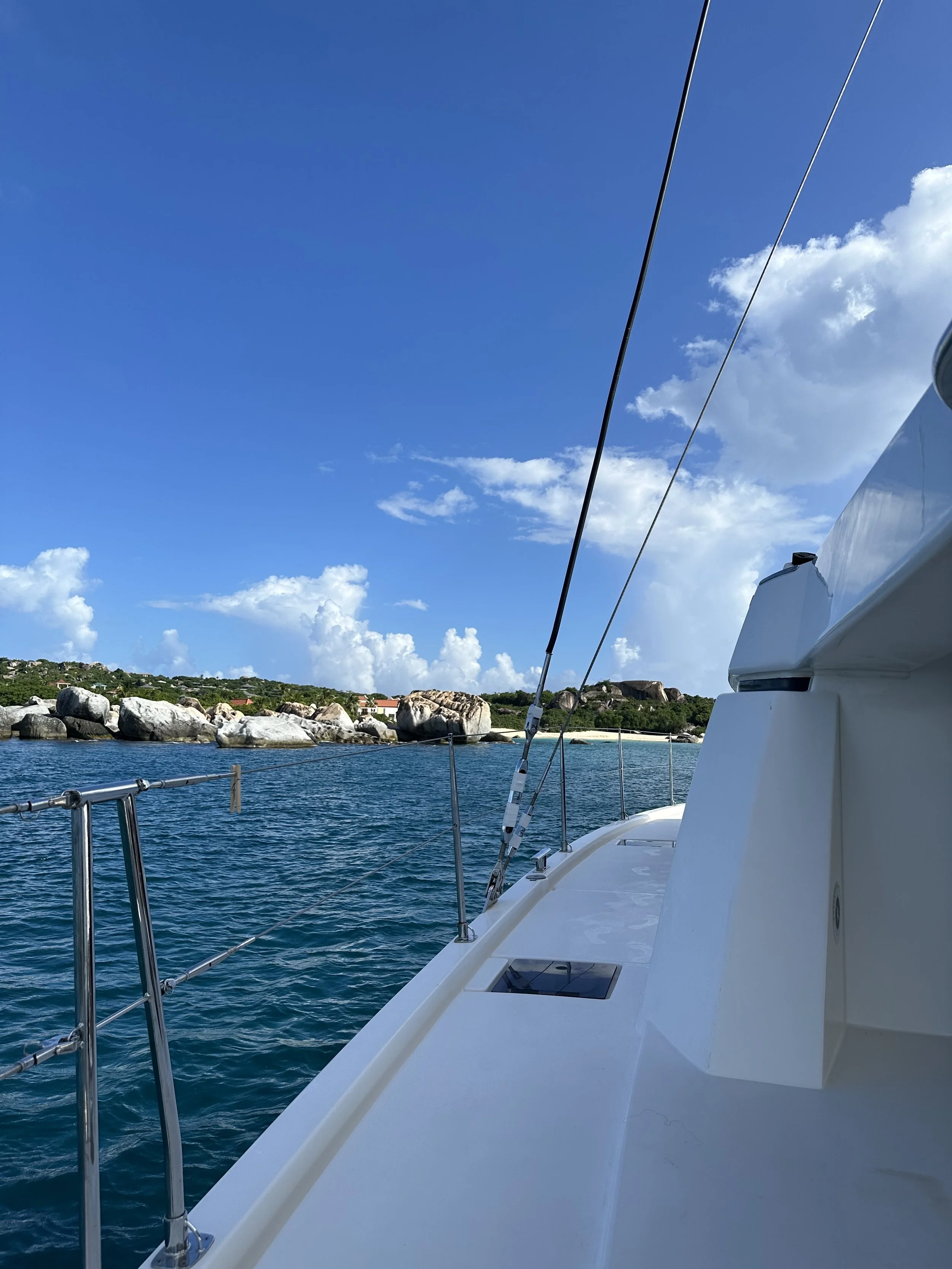 Virgin Gorda, BVI. View from a boat showing a blue sky with some clouds, the ocean water, and a rocky coastline with greenery in the background.