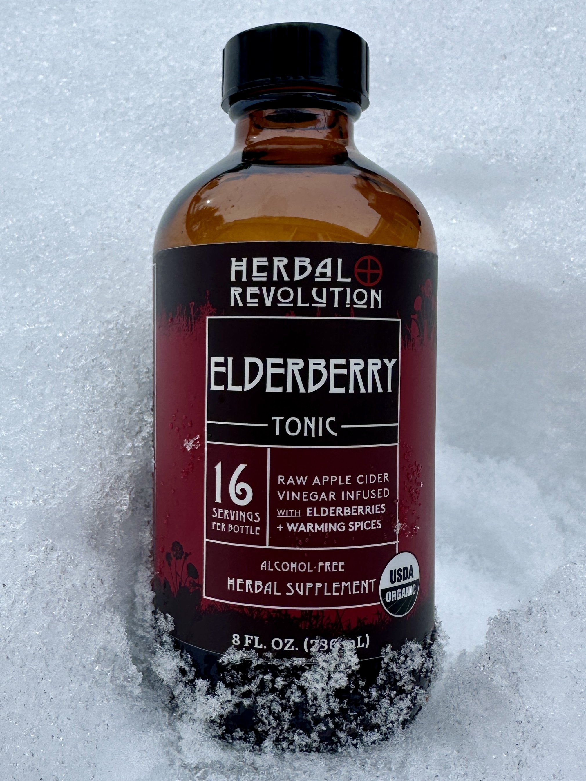 Bottle of Elderberry Tonic with elderberries and warming spices, herbal supplement, USDA organic, alcohol-free, 8 fl oz, sitting on snow.