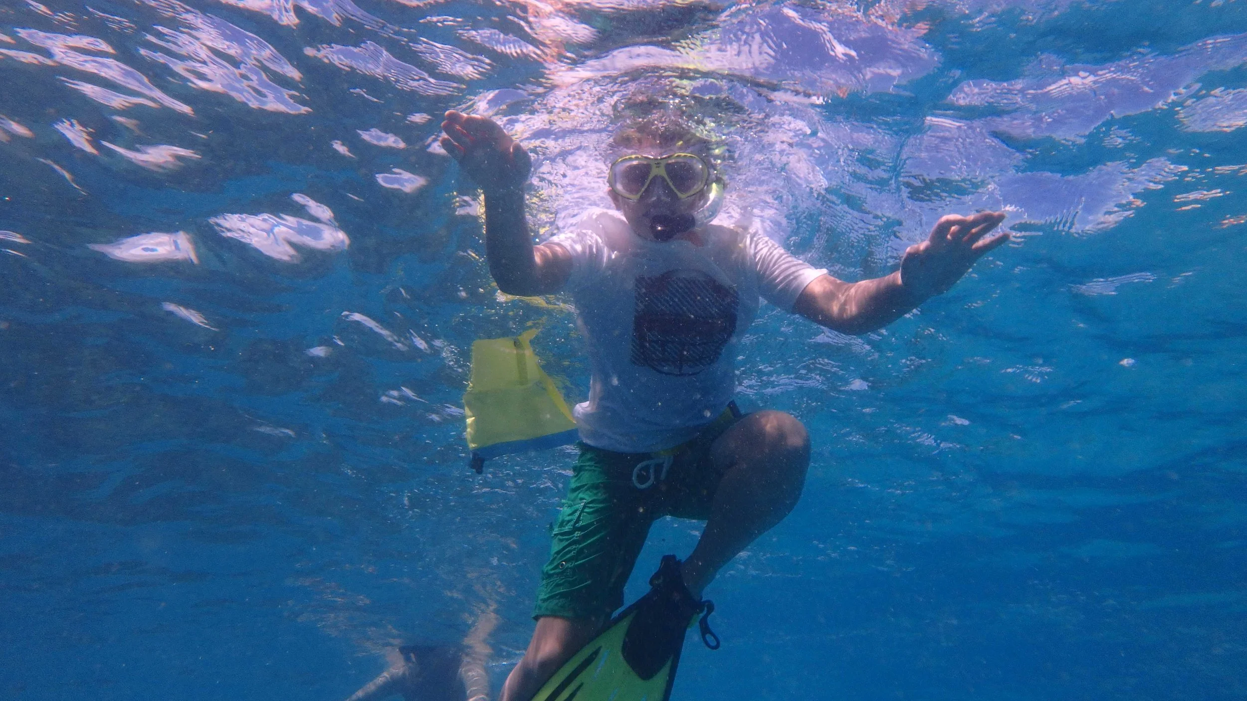 Child wearing goggles and a white t-shirt with a graphic, kneeling in shallow water, holding one hand up and extending the other while posing underwater, with waves and sunlight above.