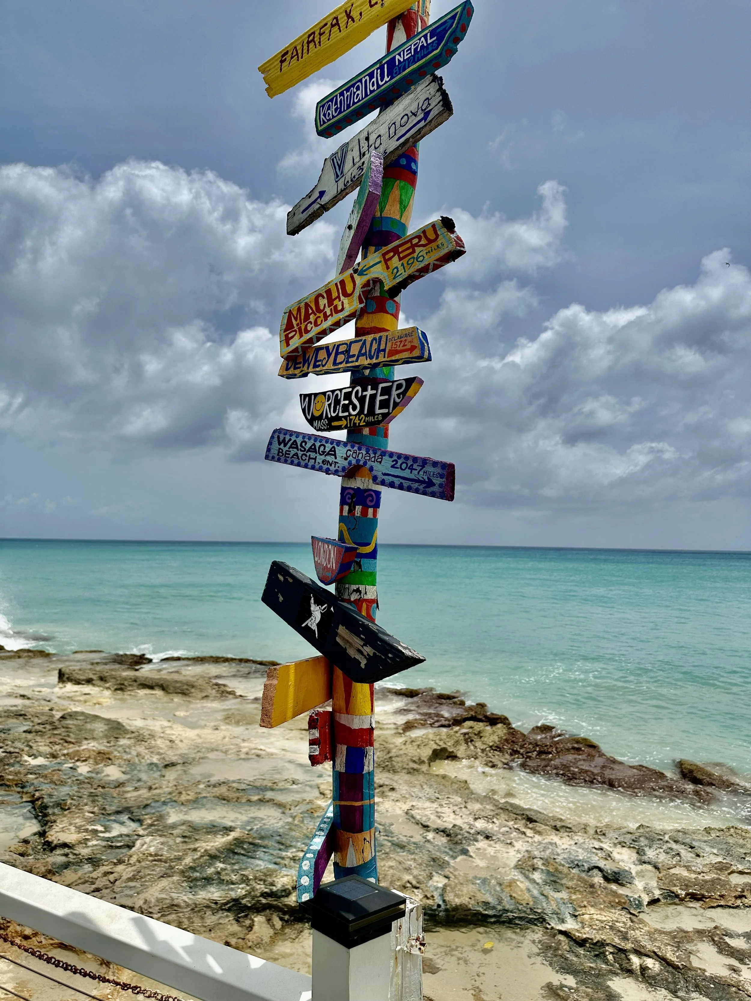 Rhythms at Rainbow Beach, St. Croix. Colorful wooden signpost with multiple directional signs pointing to various locations including Machu Picchu, London, and others, along a rocky beach with turquoise water and cloudy sky in the background.