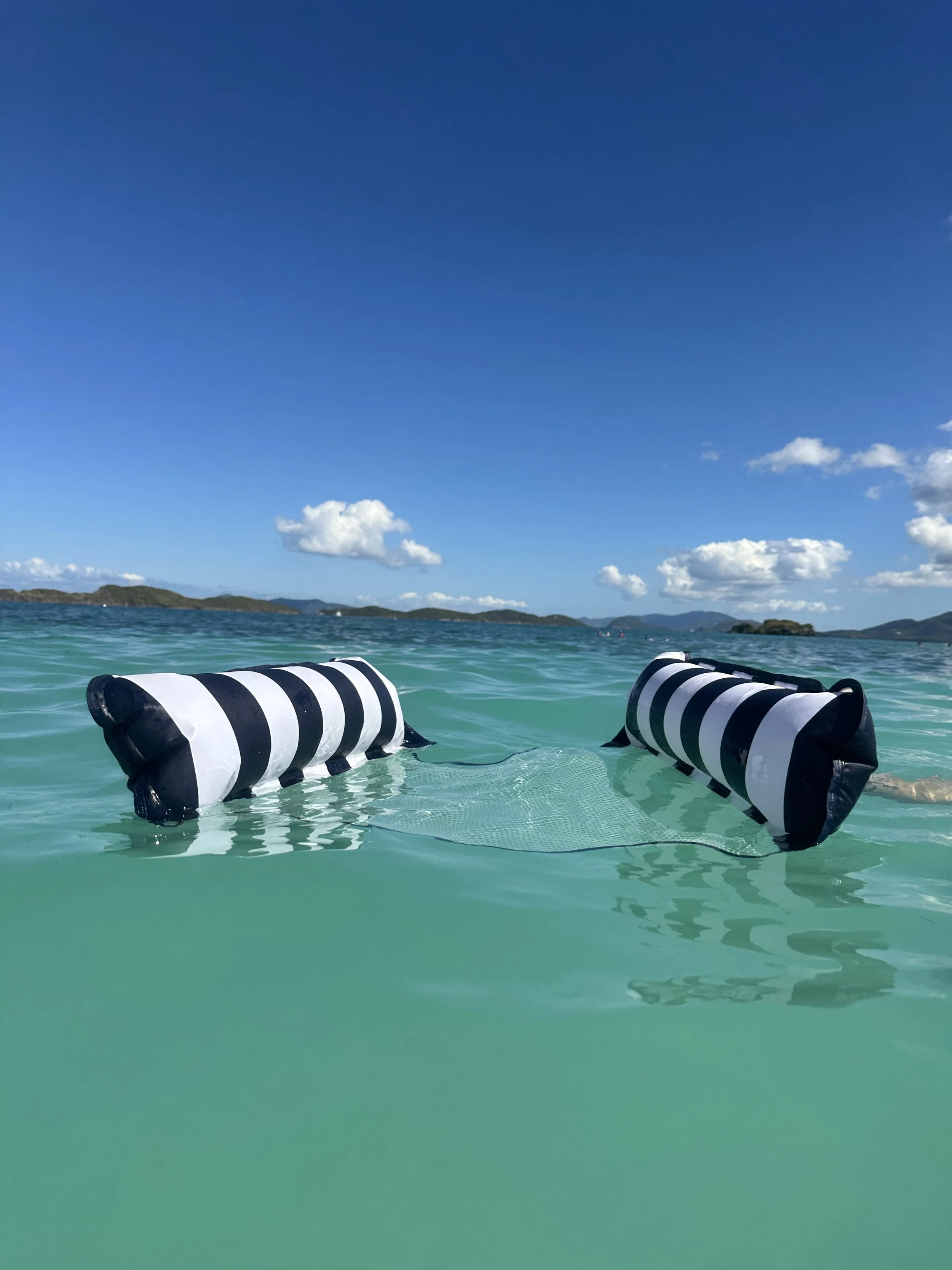 A striped black and white inflatable float in the ocean, partially submerged in clear turquoise water, with a distant island and a blue sky with scattered clouds in the background.