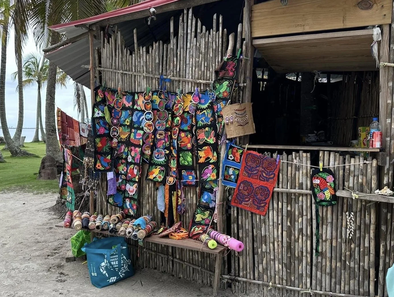 Outdoor market stall selling "mola", colorful textile bags, hats, and artwork created by the indigenous Guna people of San Blas islands Panama, hanging on a bamboo fence, with palm trees and a grassy area in the background.