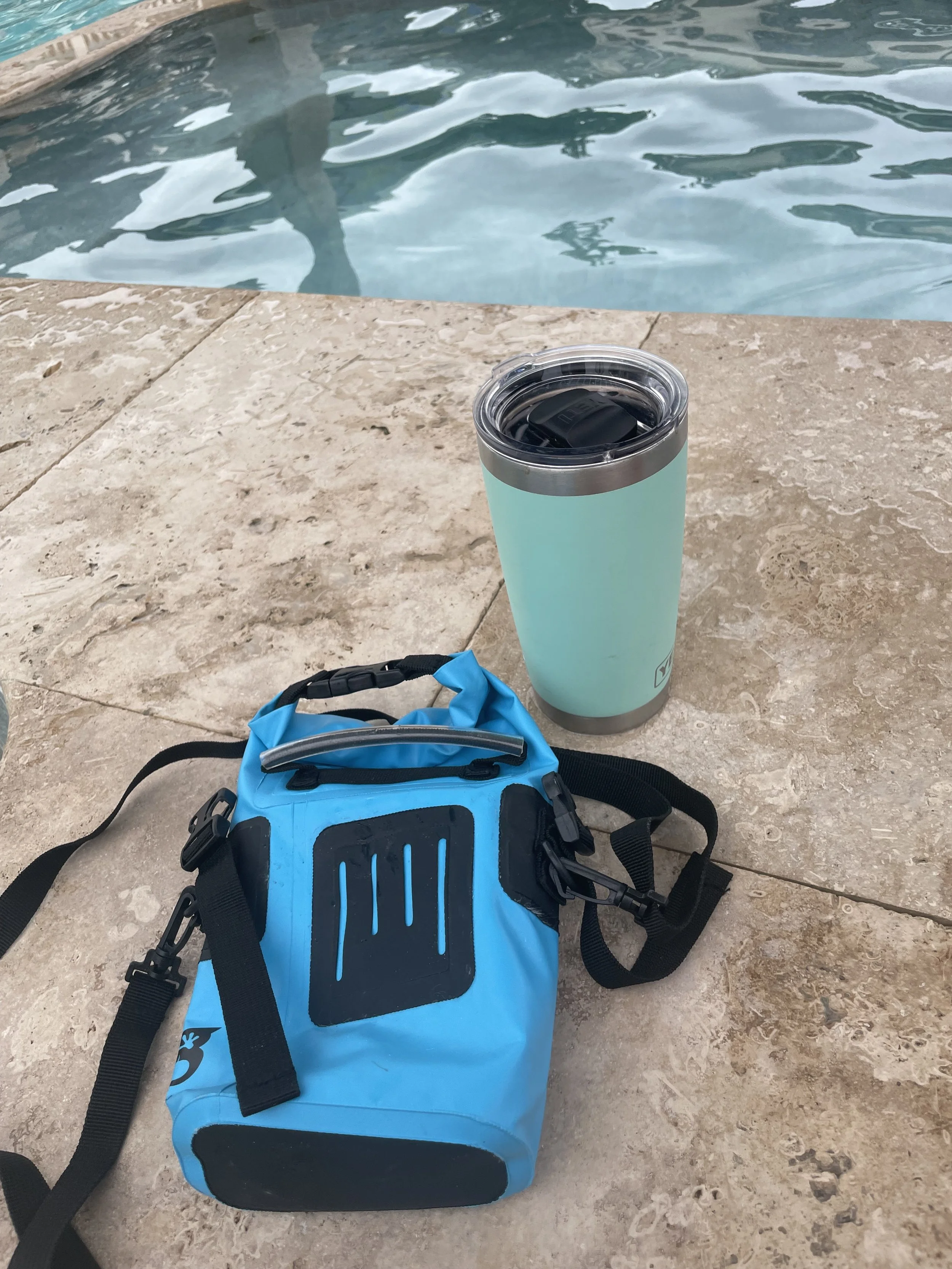 A blue waterproof bag and a light blue tumbler with a black lid are placed on a tan stone poolside surface. The background shows a swimming pool with clear water.