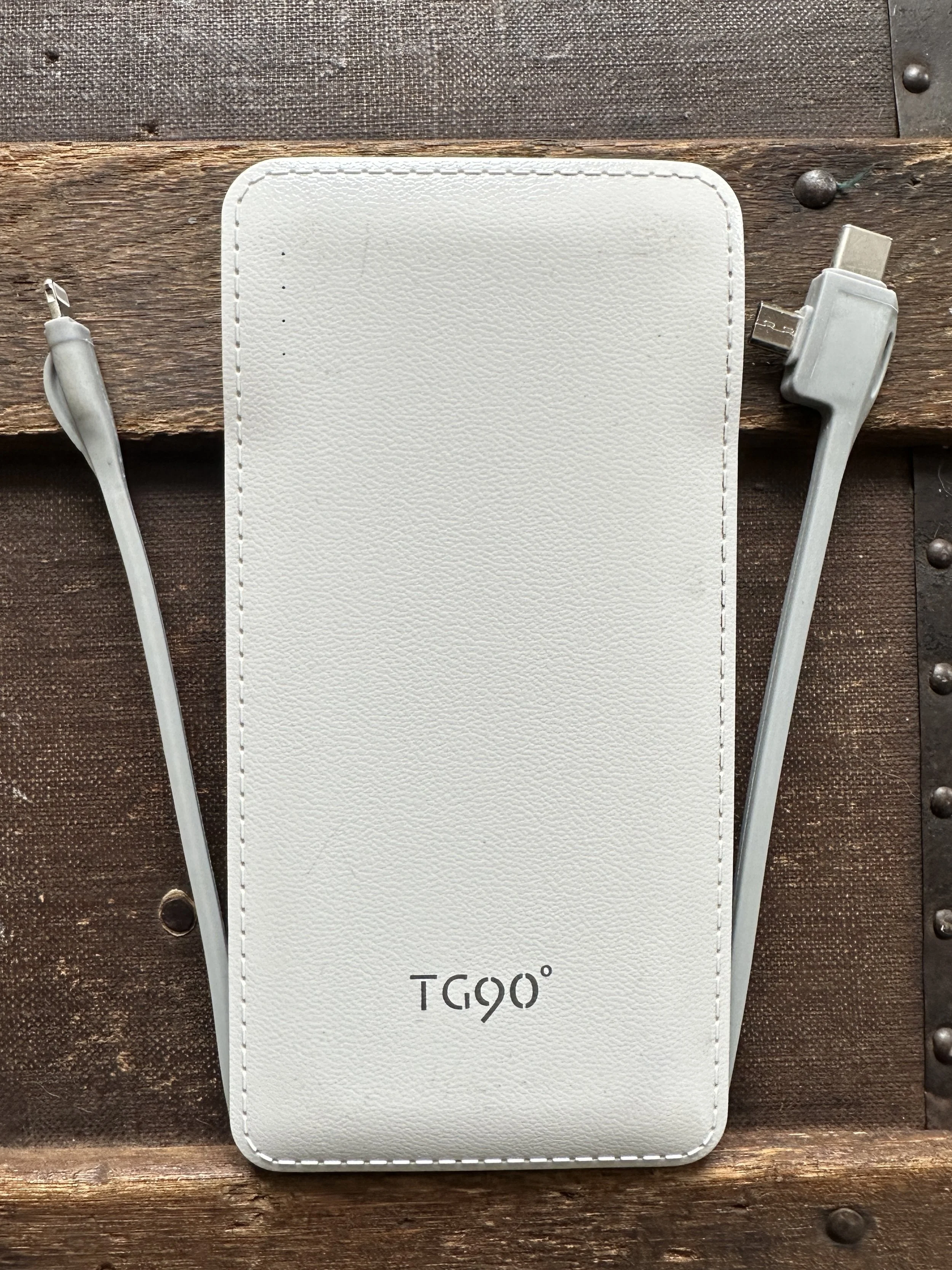 A white portable handheld fan with the brand name "Tego" printed on it, placed on a wooden surface with a charger and cable nearby.
