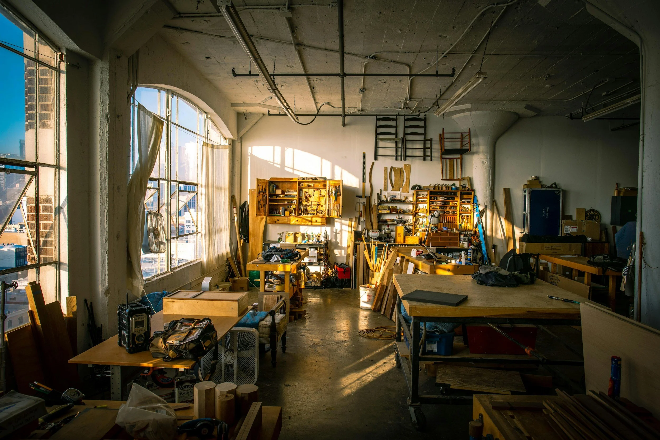A woodworking workshop with sunlight streaming through large windows, with various tools, wood pieces, and workbenches visible.