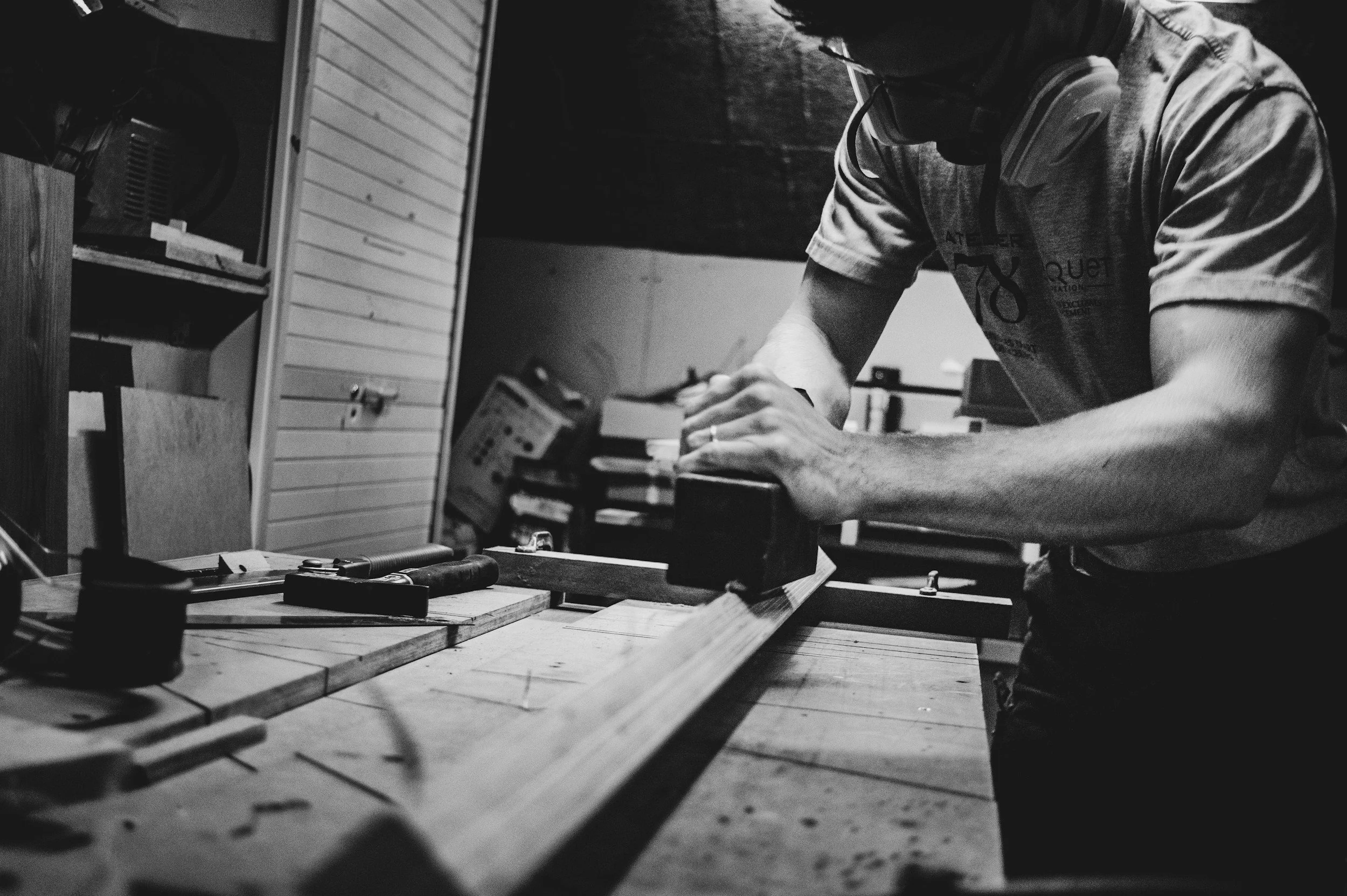 A person working on a woodworking project, using tools and clamps on a wooden workbench inside a workshop.