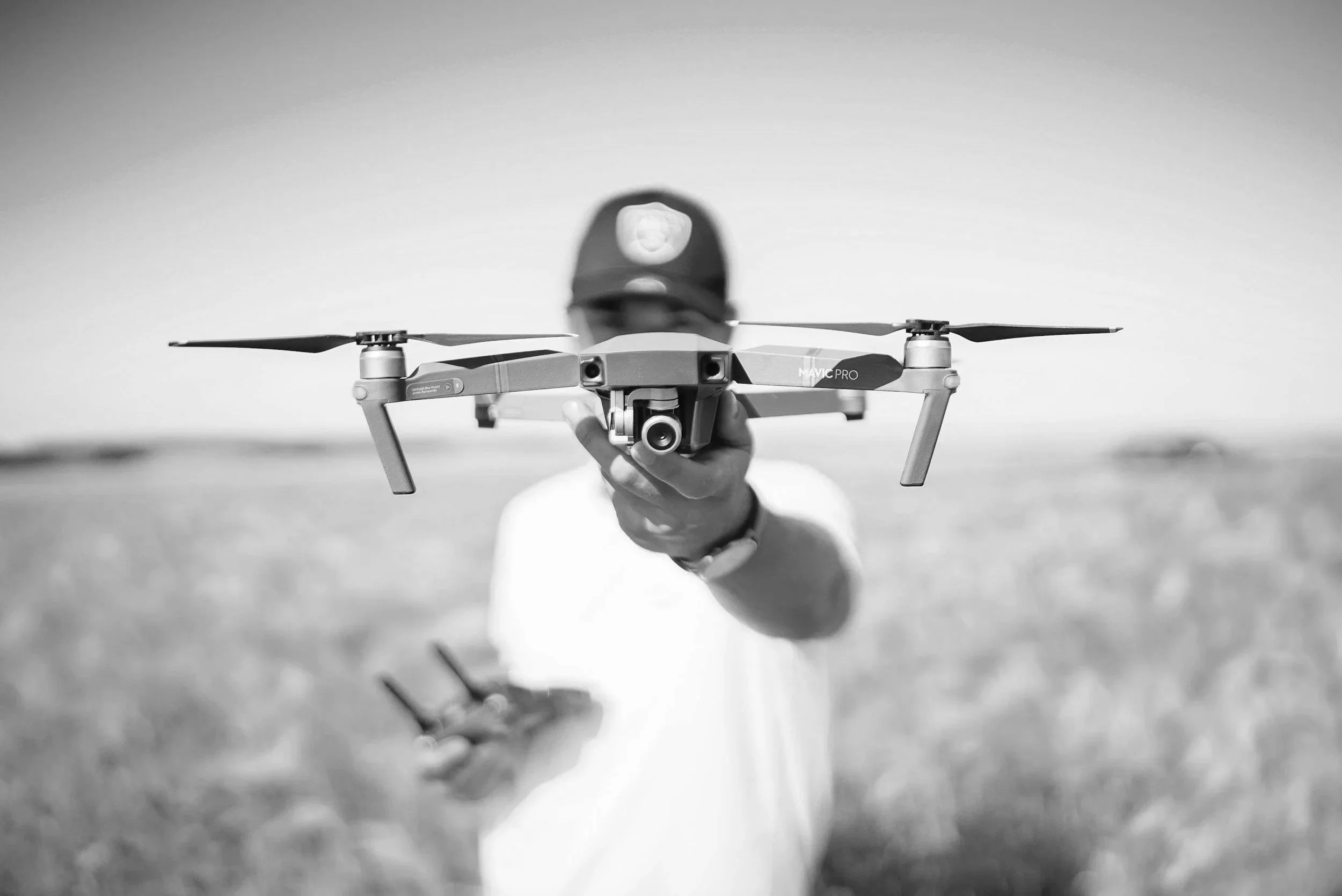 A person wearing a cap and white shirt aiming a drone with a camera attached, holding a remote control in an open field.