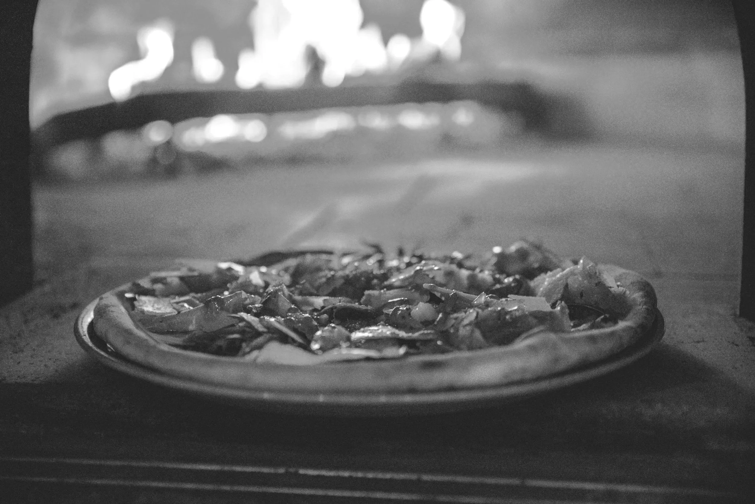 A pizza baking inside a pizza oven, seen from the front in black and white.
