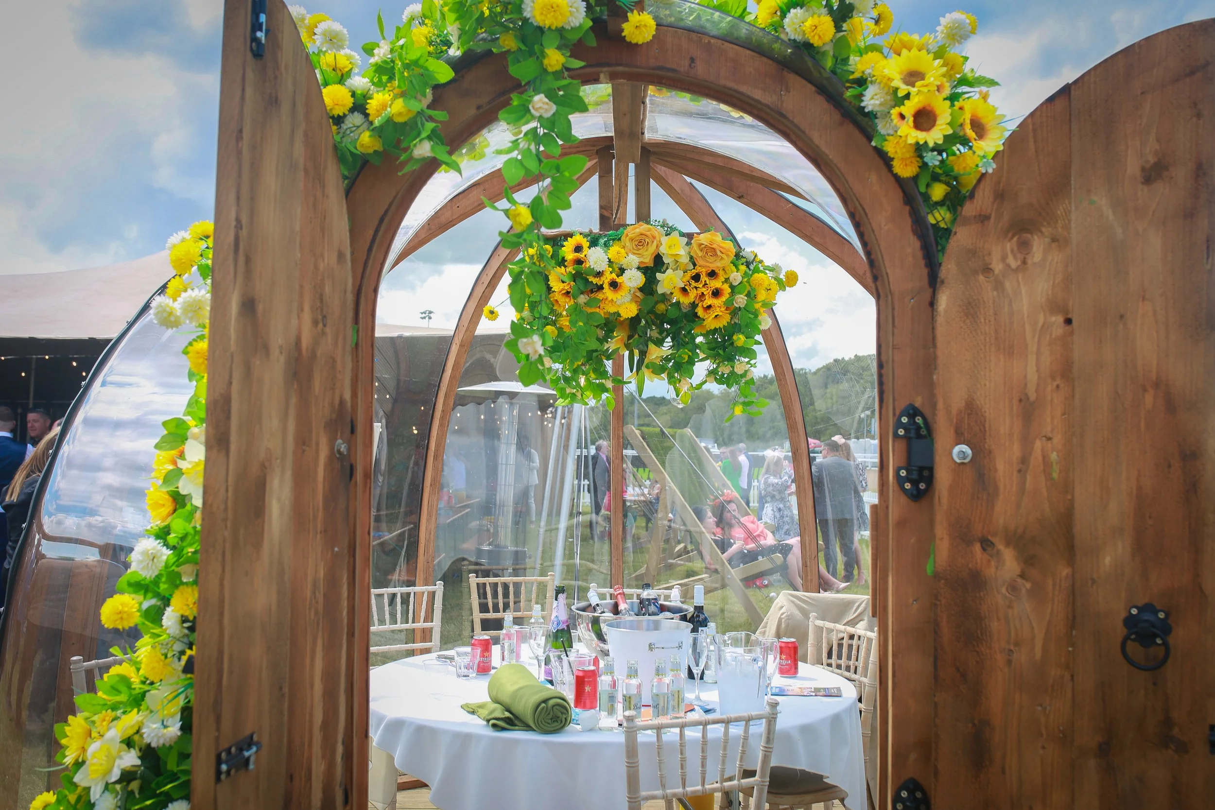 View through a wooden and glass door of a decorated outdoor event space with a table set for dining, featuring a white tablecloth, bottles, glasses, and a rolled green napkin, with floral arrangements of yellow and white flowers hanging from the ceiling, and people and tents visible in the background.
