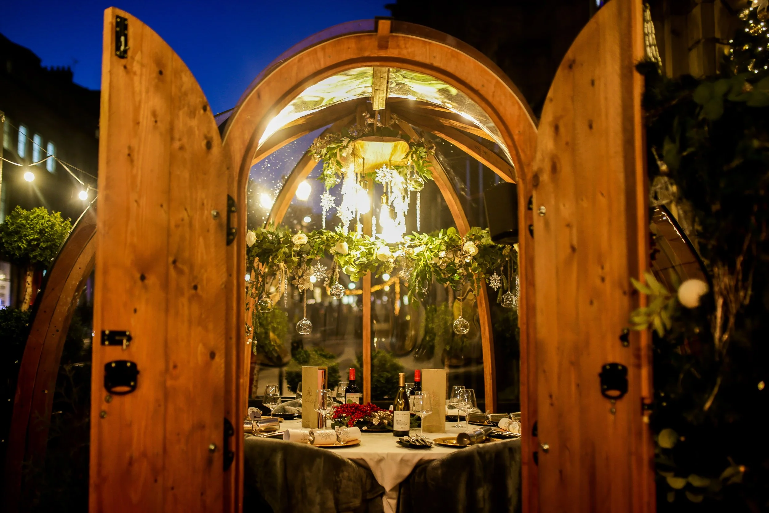 A cozy dining setup inside a wooden structure with a table set for a meal, decorated with holiday ornaments, hanging glass globes, greenery, and string lights outdoors at night.