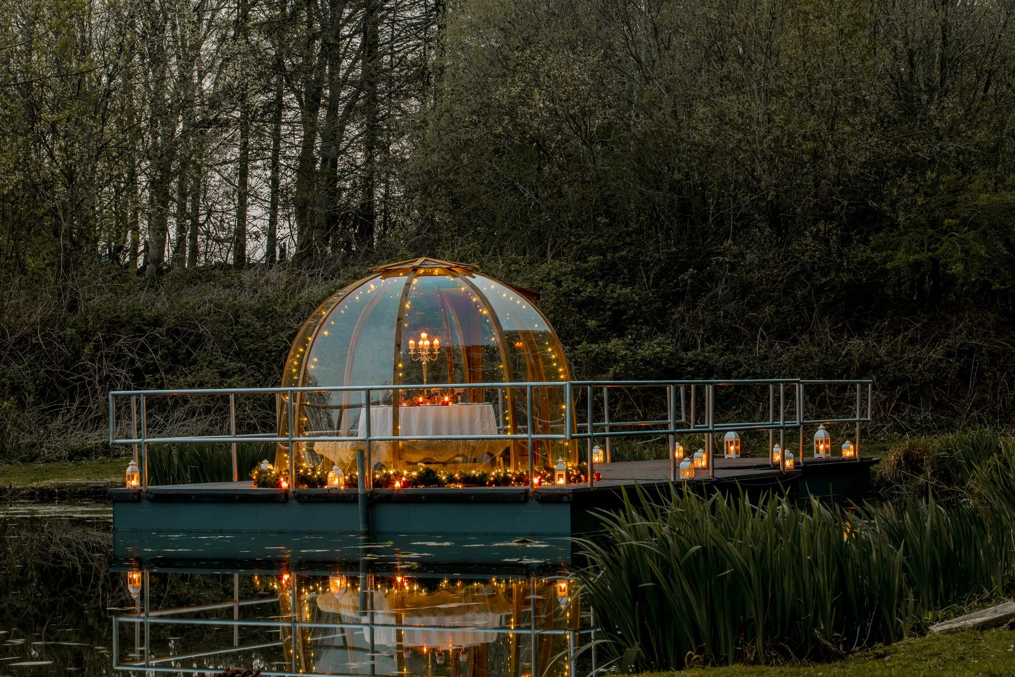 A small glass dome on a floating platform over water, decorated with lanterns and string lights, with a table inside set with candles and a candelabra, surrounded by lush greenery and trees.