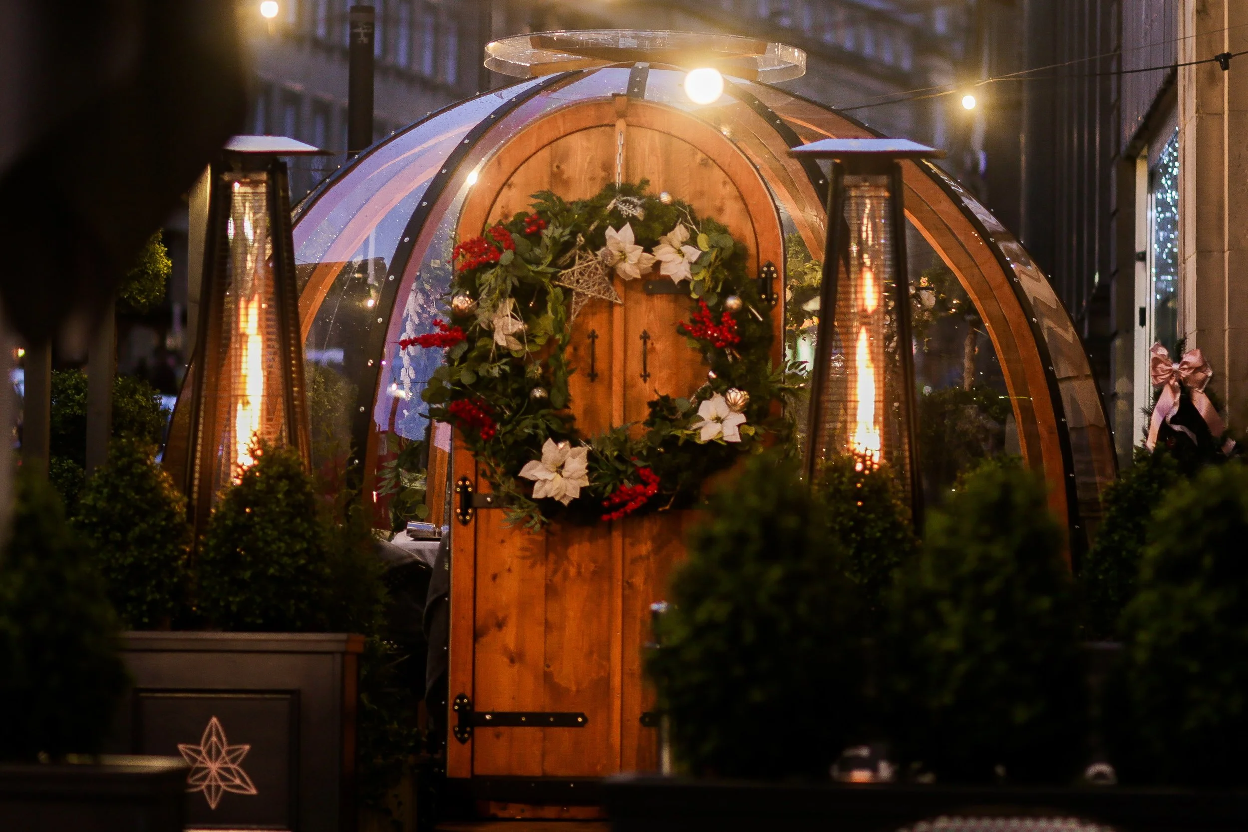 A small, rounded wooden structure decorated for Christmas with a wreath of poinsettias, holly, and red berries on the door, surrounded by outdoor heaters and greenery at night.