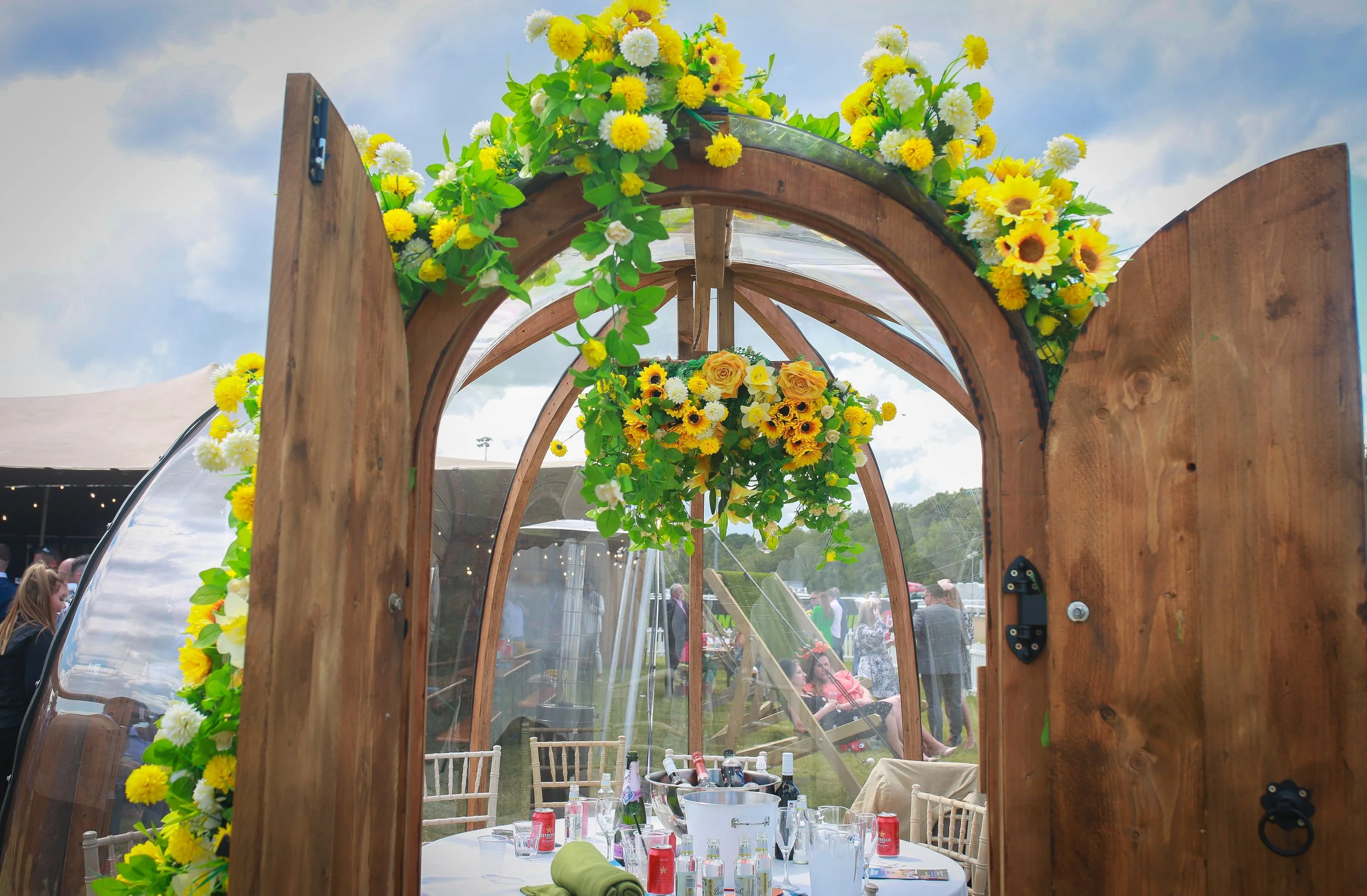 Wooden wedding or event archway decorated with yellow and white flowers, leading to a table set with beverages and glassware, outdoors with people and tents in the background.